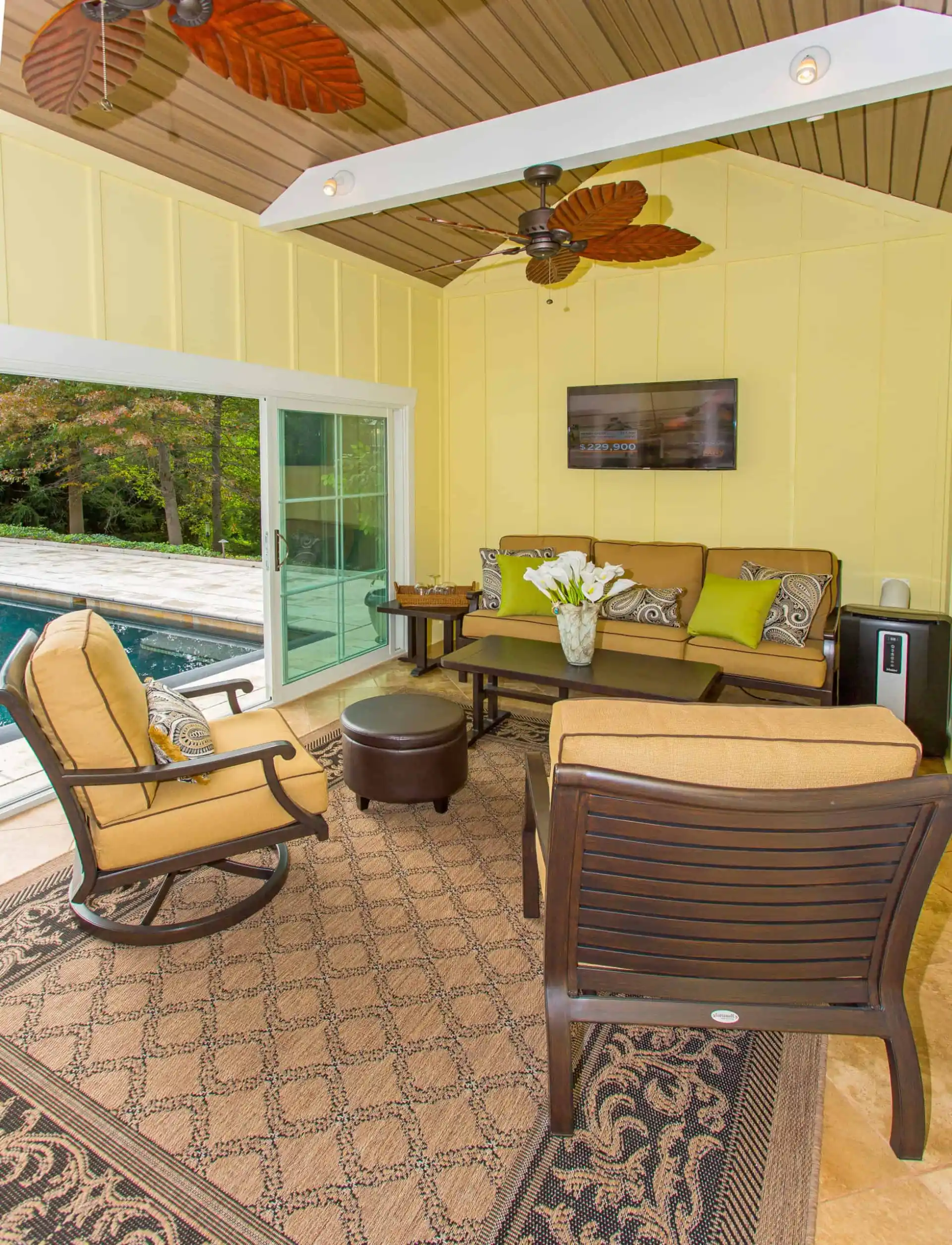 A cozy sunroom with cushioned chairs, a sofa with green pillows, a patterned rug, a coffee table with white flowers, a wall-mounted TV, ceiling fans, and large glass doors opening to a pool area outside.