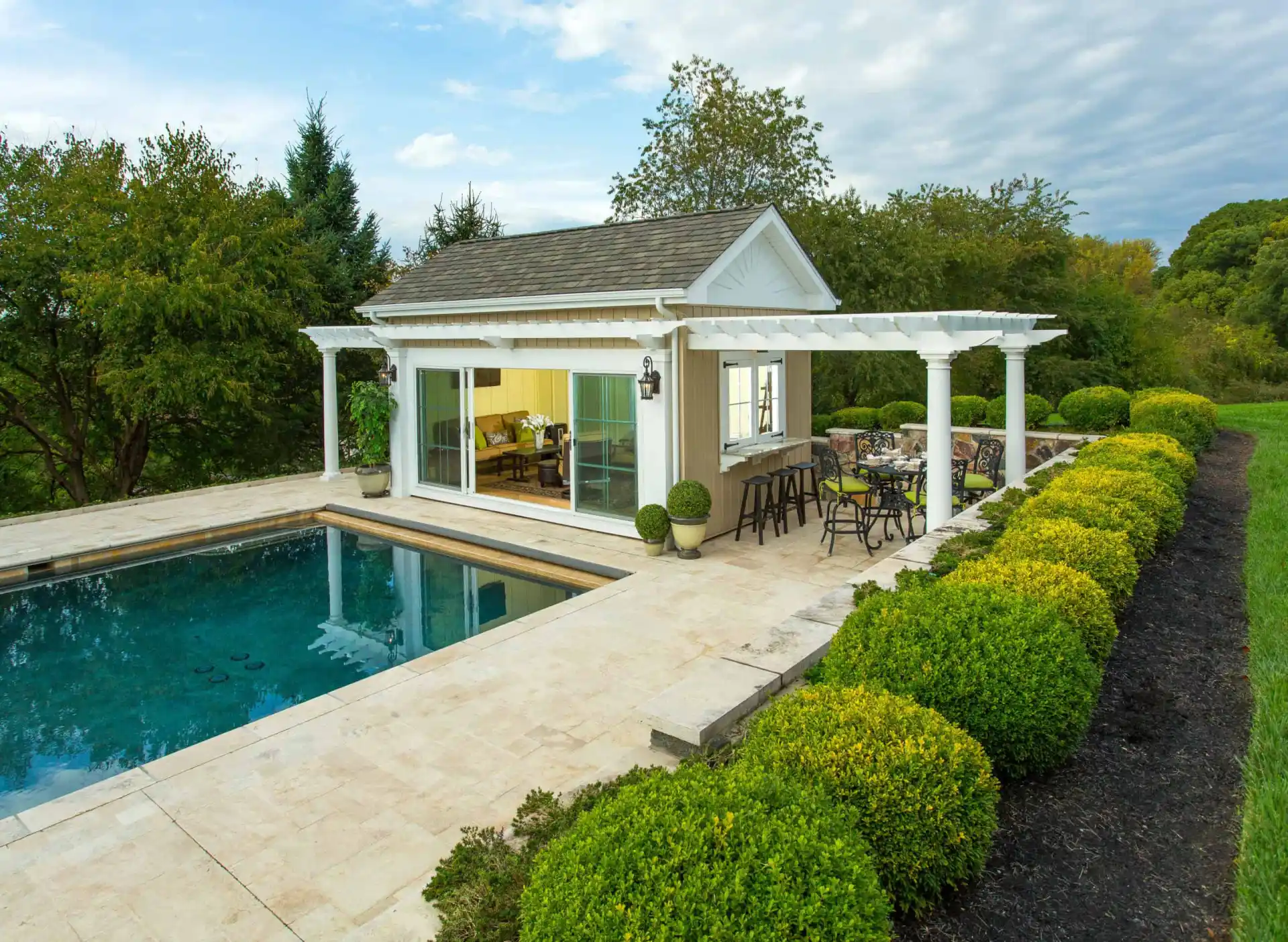 A small pool house with white columns and a pergola sits beside a rectangular pool, surrounded by manicured bushes and trees. Glass doors reveal a cozy interior, and outdoor seating lines the patio under a partly cloudy sky.