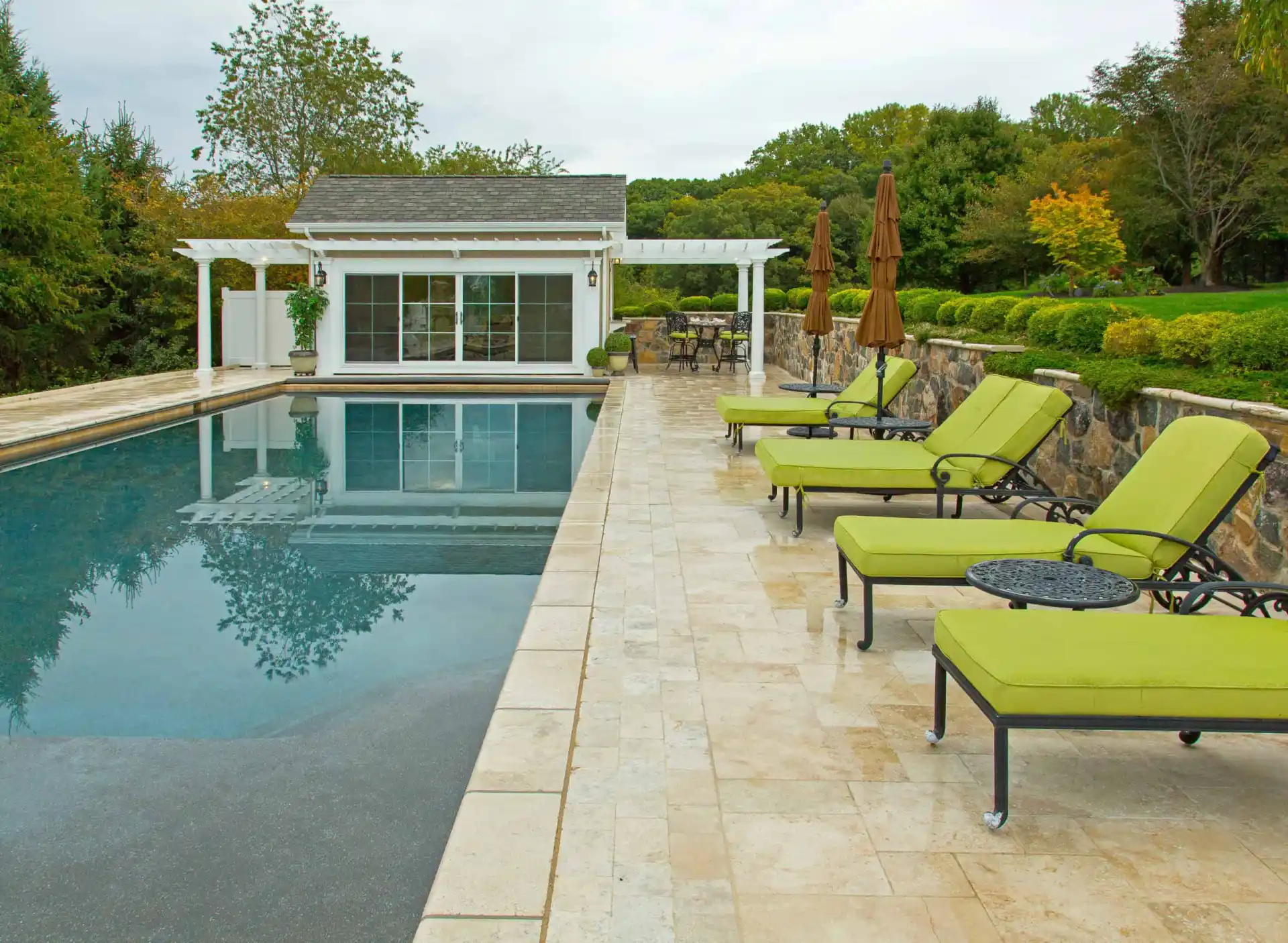 A backyard pool with a stone deck, several green cushioned lounge chairs, round tables, brown umbrellas, and a pool house with large windows surrounded by greenery and trees.