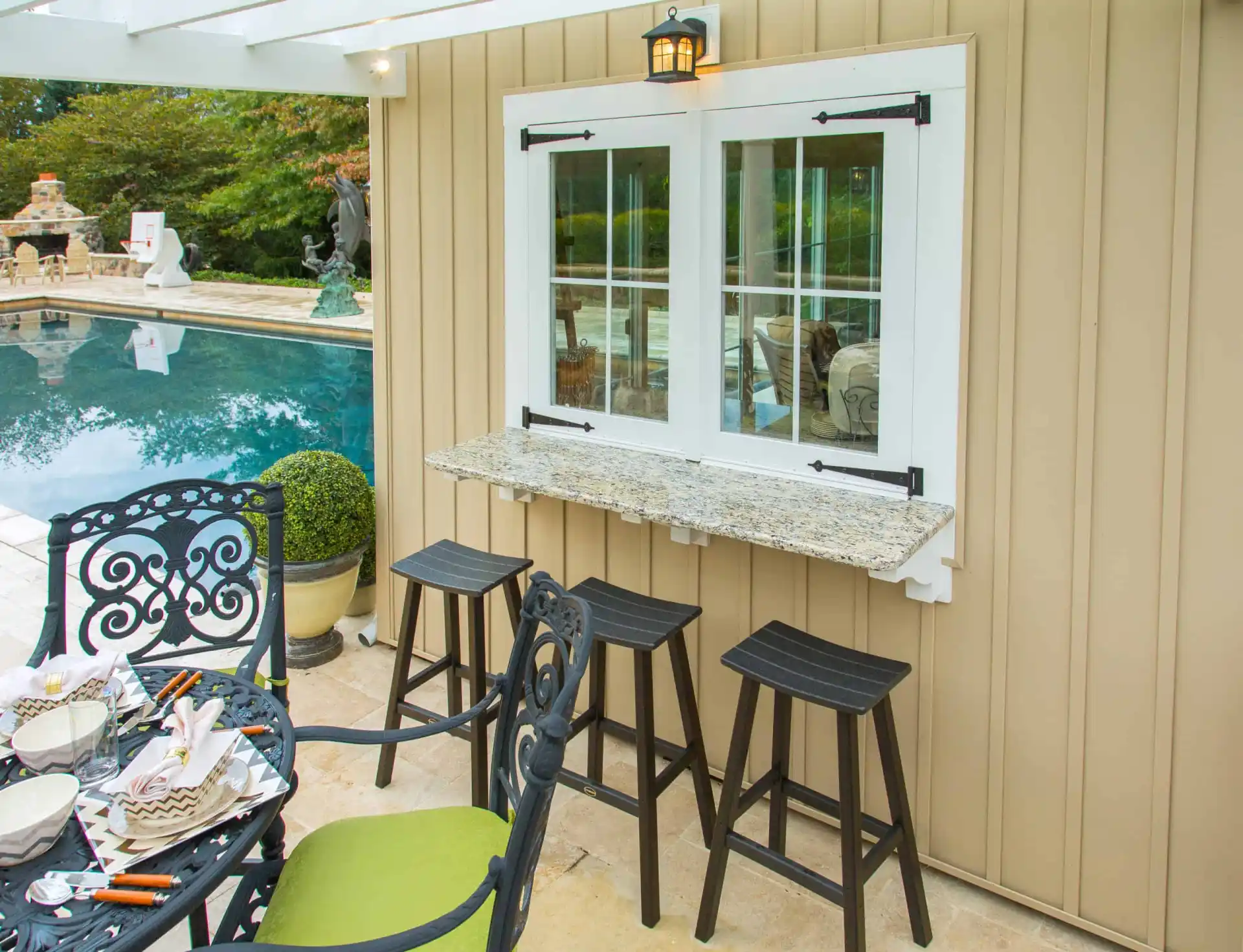 Outdoor patio area with a granite bar ledge beneath a window, three black stools, decorative metal chairs, and a table next to a pool. Lush greenery and a stone fireplace are visible in the background.