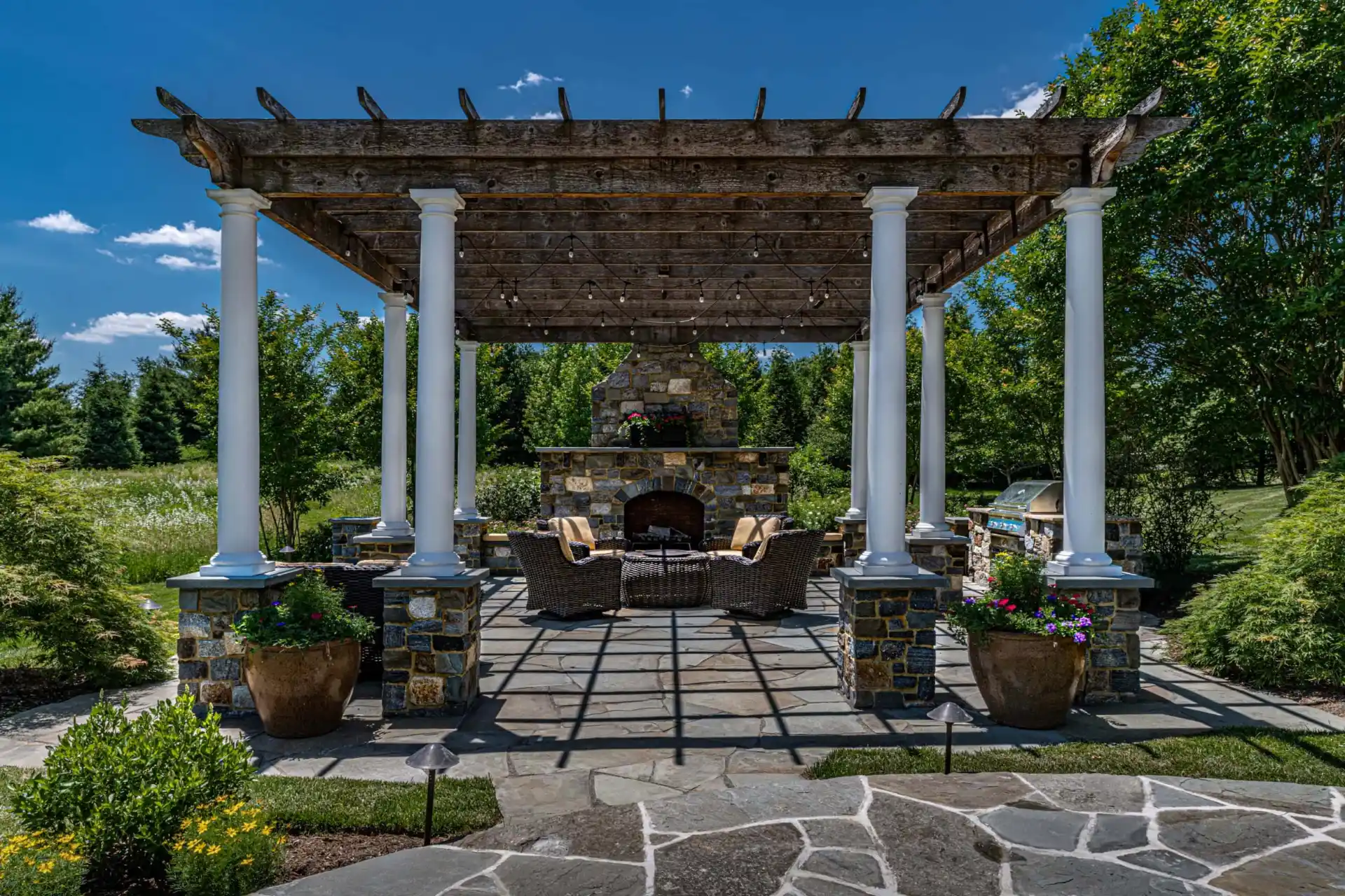 A rustic outdoor patio with a stone fireplace under a wooden pergola offers the perfect setting for outdoor living in Delaware County, PA. Wicker seating surrounds the fireplace, while potted plants and expert hardscaping services complete this serene scene against a backdrop of trees and blue sky.