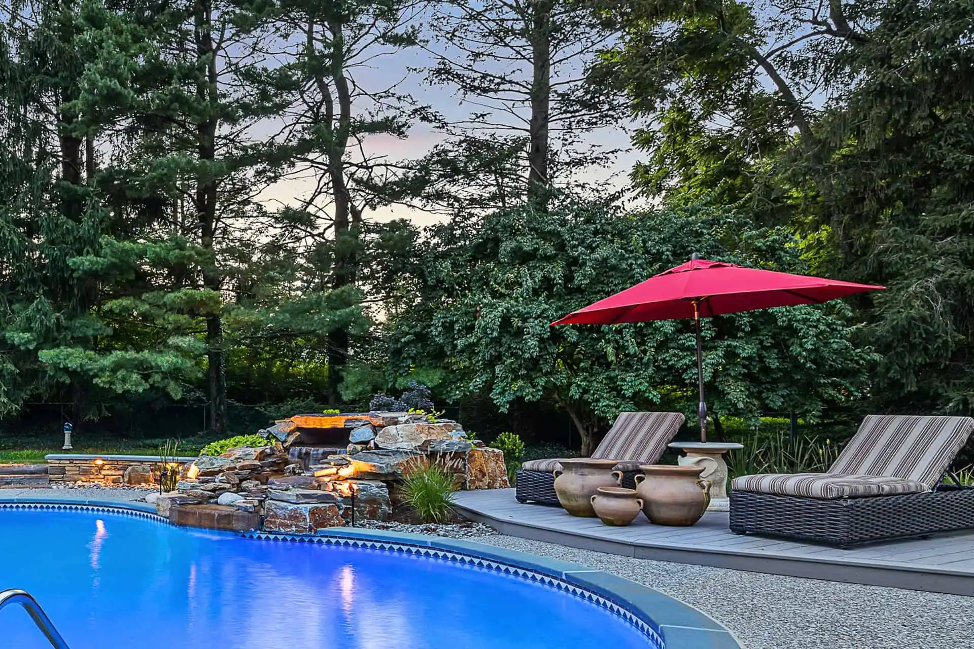 A backyard pool with glowing blue water, surrounded by trees. A stone waterfall feature, two striped lounge chairs, clay pots, and a red umbrella sit on the pool deck.