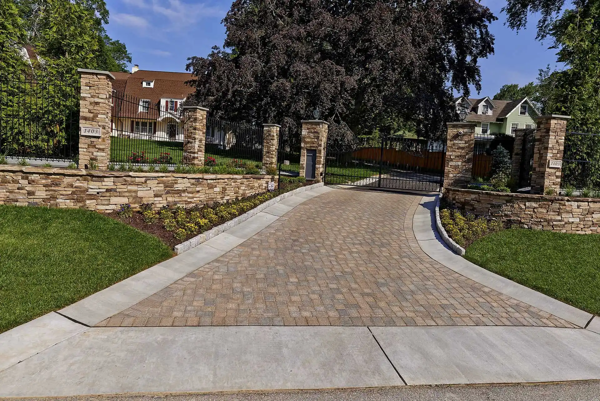A wide stone-paved driveway leads to a closed iron gate with brick pillars, surrounded by manicured lawns, shrubs, and trees, with residential houses visible in the background.