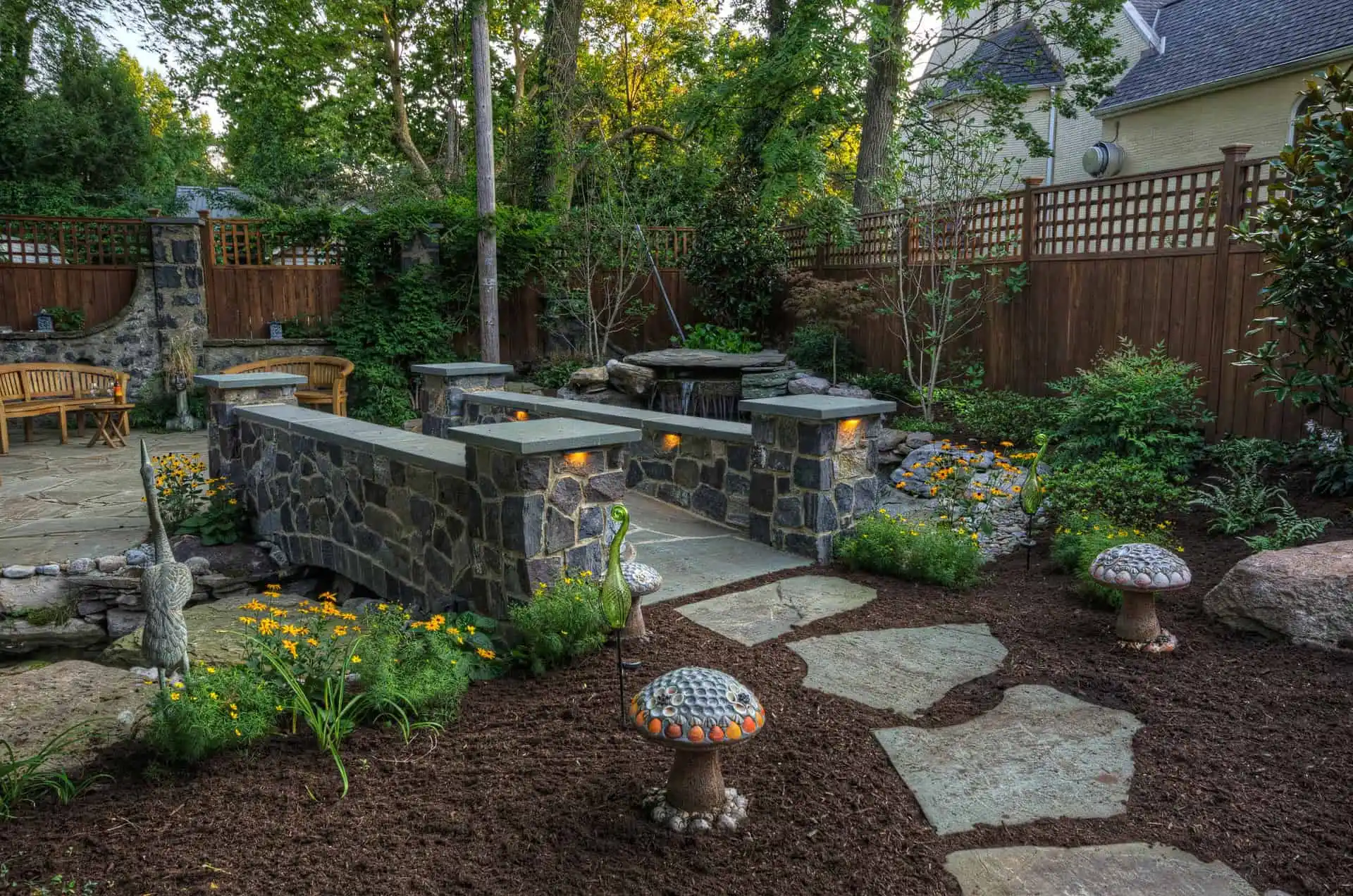A landscaped backyard garden with stone paths, decorative mushroom sculptures, flower beds, and a stone bridge with built-in lights, surrounded by lush greenery and wooden fencing.