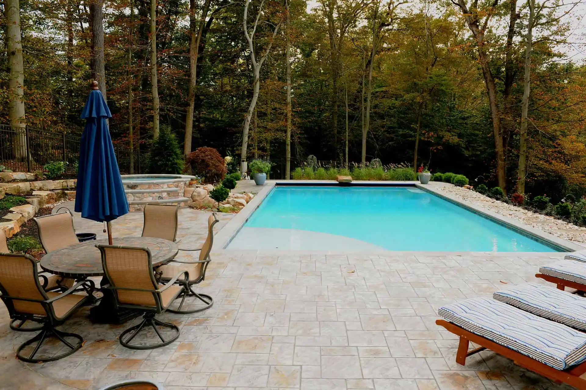 A backyard with a rectangular swimming pool, surrounded by trees. There’s a patio area with cushioned lounge chairs and a round table with chairs and a closed blue umbrella. The scene appears peaceful and inviting.