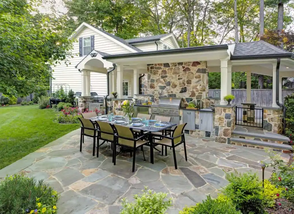 Outdoor patio with a dining table set for eight on a stone floor, an inviting space that showcases the essence of outdoor living in Delaware County, PA. A built-in grill and countertop are under a covered area with stone walls, while lush greenery and flowers adorn the landscaped yard beside a partially visible white house.