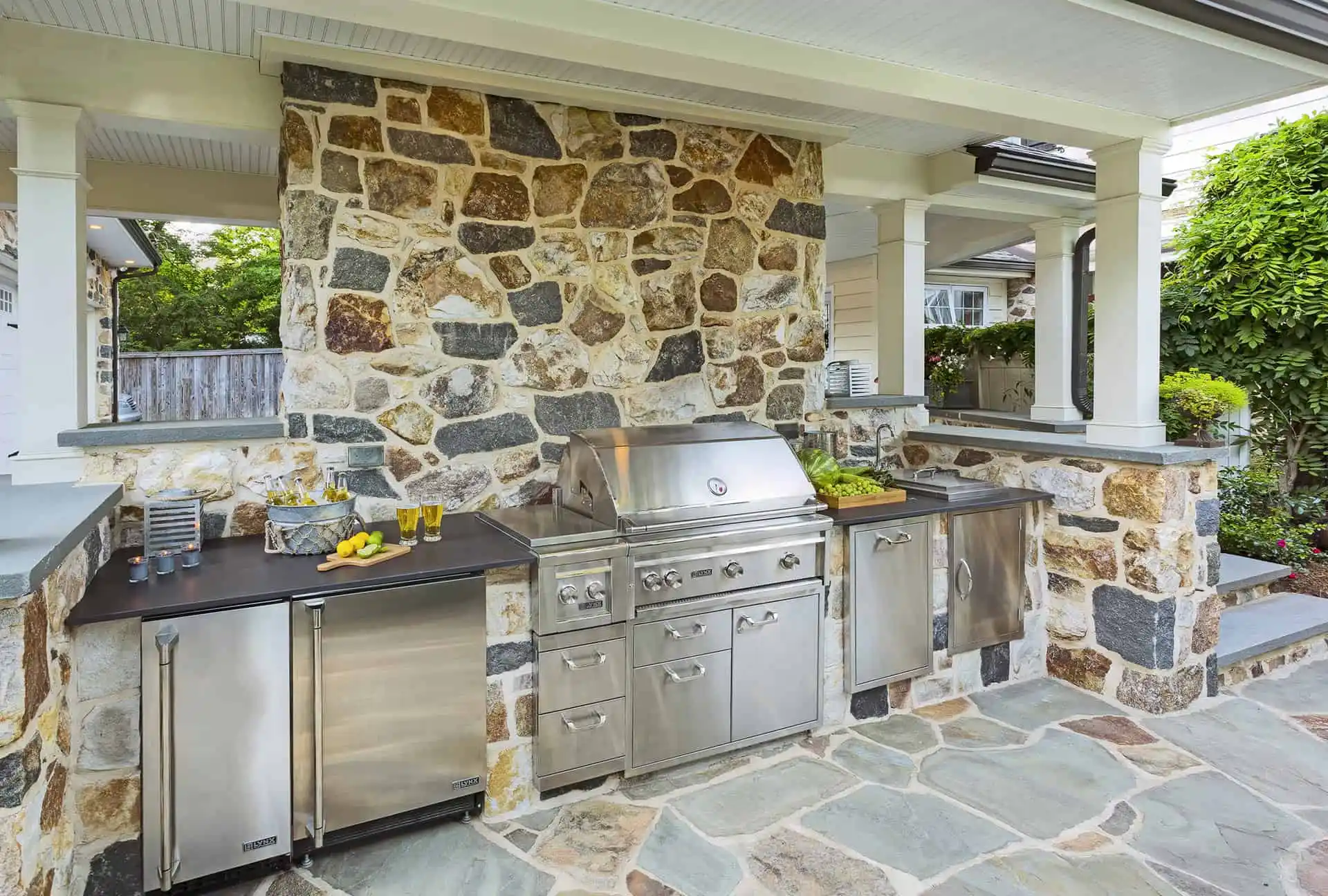 Outdoor kitchen with a built-in stainless steel grill, refrigerator, and sink set against a stone wall under a covered patio, with a stone tile floor and green plants visible in the background.