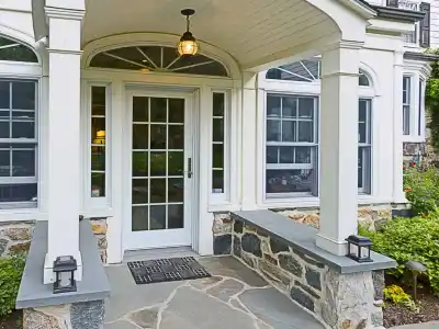 A front entrance of a house with a stone porch and archway showcases expert hardscaping services in Delaware County, PA. The door is surrounded by large windows, with two lanterns resting on the stone walls flanking the walkway. The exterior blends stone and siding, enriched by lush greenery.