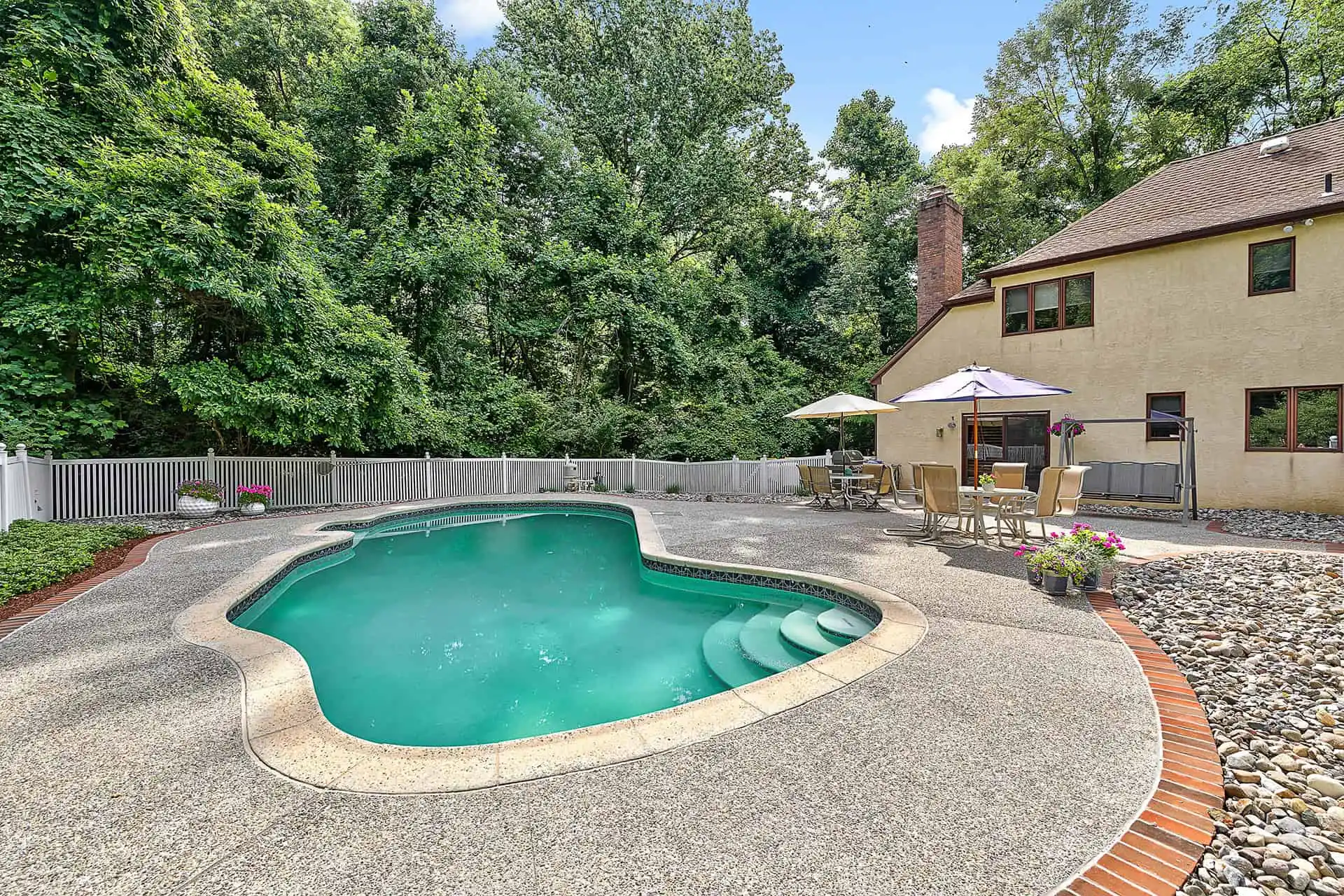 Backyard with an in-ground swimming pool, surrounded by a patio area with outdoor dining furniture, umbrellas, potted plants, and a beige house; trees and a white fence in the background.