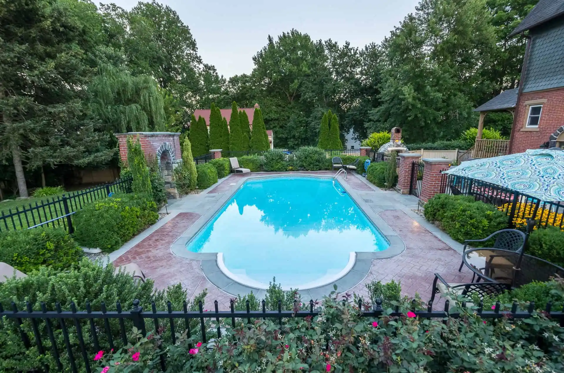 A rectangular outdoor swimming pool surrounded by brick paving, greenery, lounge chairs, and a decorative fence, with trees and a house in the background on a clear day.