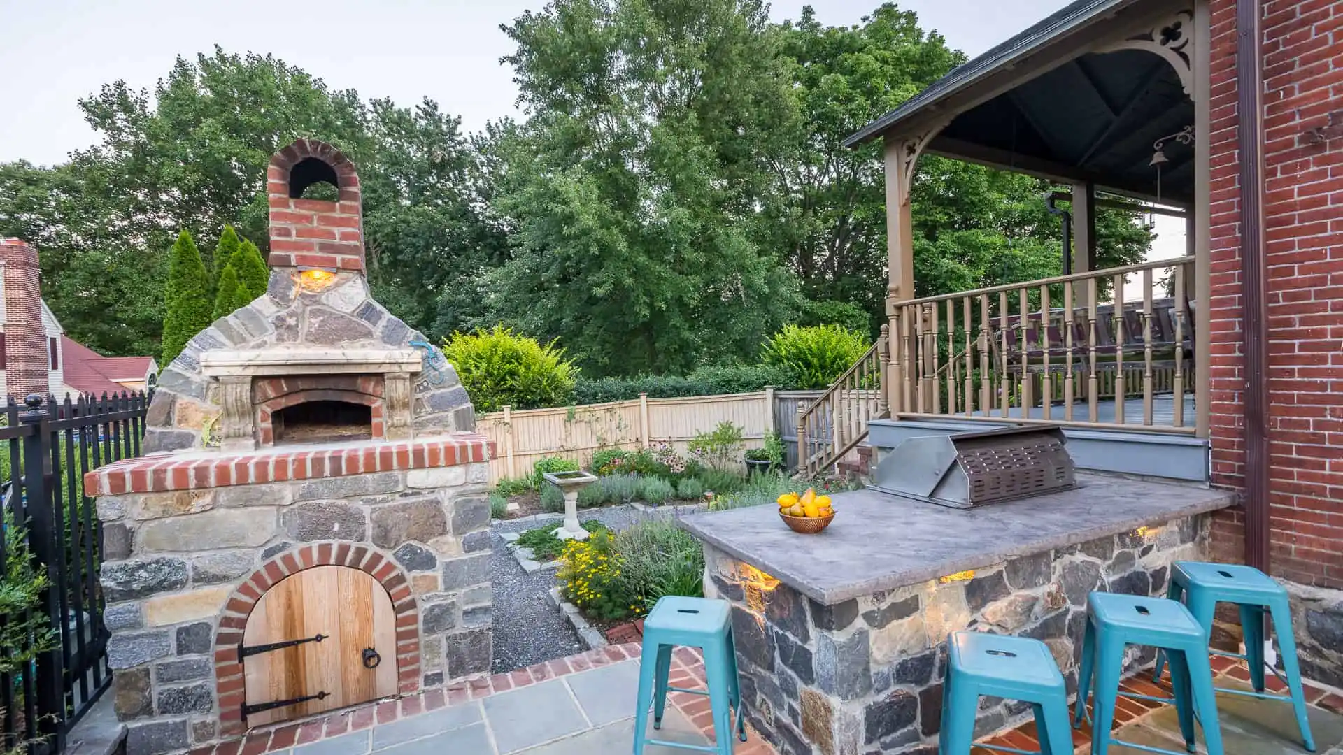 Outdoor patio with a stone brick pizza oven, adjacent countertop with four blue stools, and a bowl of oranges. Behind is a wooden fence, lush greenery, and part of a brick house with a covered porch—perfect for those enjoying outdoor living in Delaware County, PA.