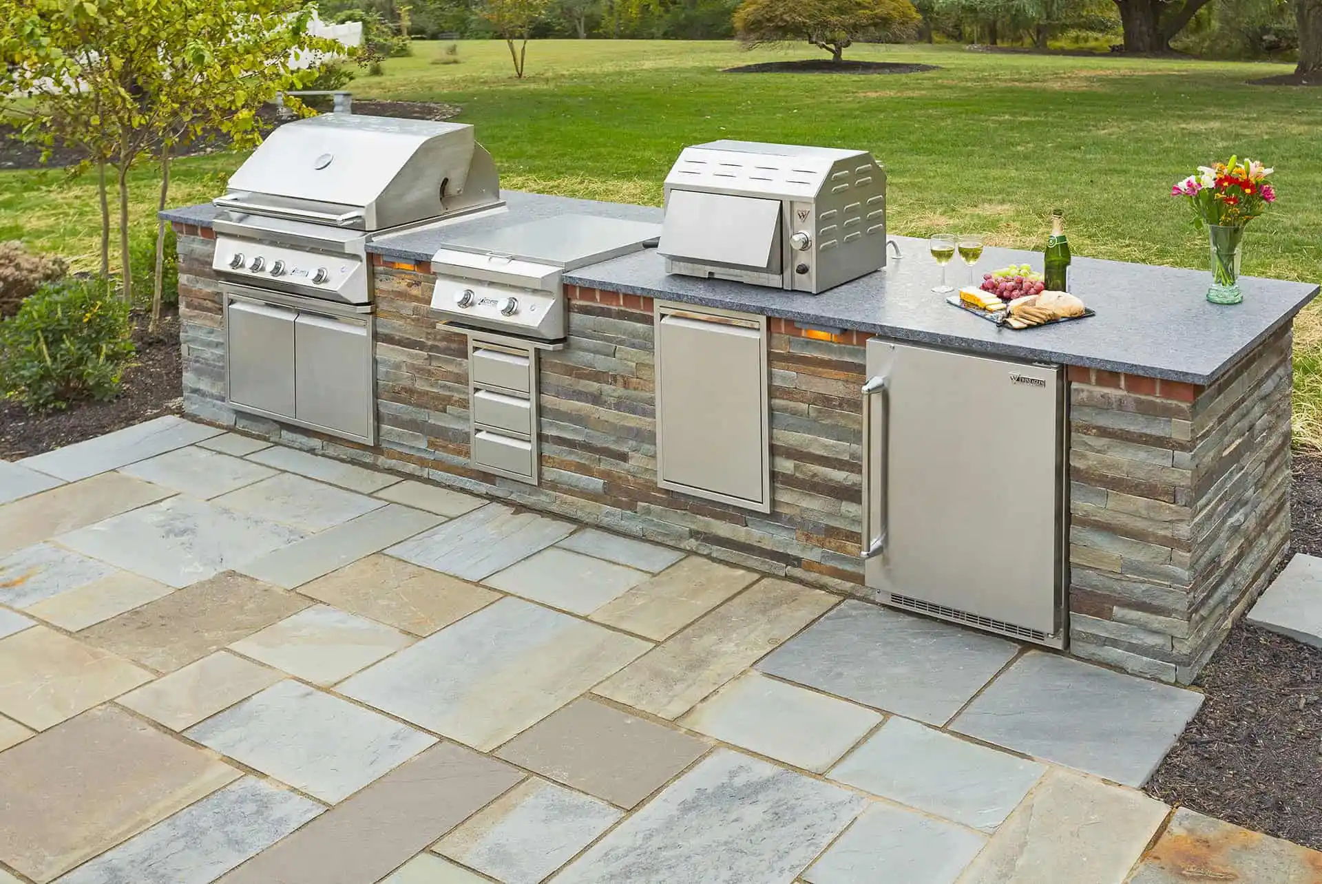 A modern outdoor kitchen with stainless steel appliances, including grills, a refrigerator, and a prep area with wine glasses, a bouquet of flowers, and food on a stone patio overlooking a green yard.