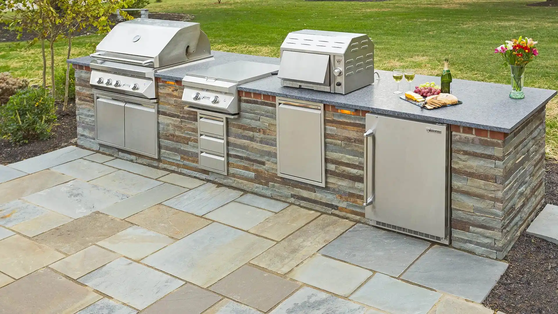 Outdoor living in Delaware County, PA is elevated with a stone countertop kitchen, featuring a built-in grill, side burner, and refrigerator. Enjoy bread on the cutting board and wine by the flower vase. Stone pavers complete this charming setup of hardscaping services in Delaware County, PA.