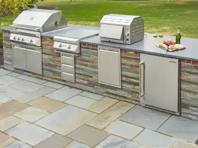 Outdoor living in Delaware County, PA is elevated with a stone countertop kitchen, featuring a built-in grill, side burner, and refrigerator. Enjoy bread on the cutting board and wine by the flower vase. Stone pavers complete this charming setup of hardscaping services in Delaware County, PA.