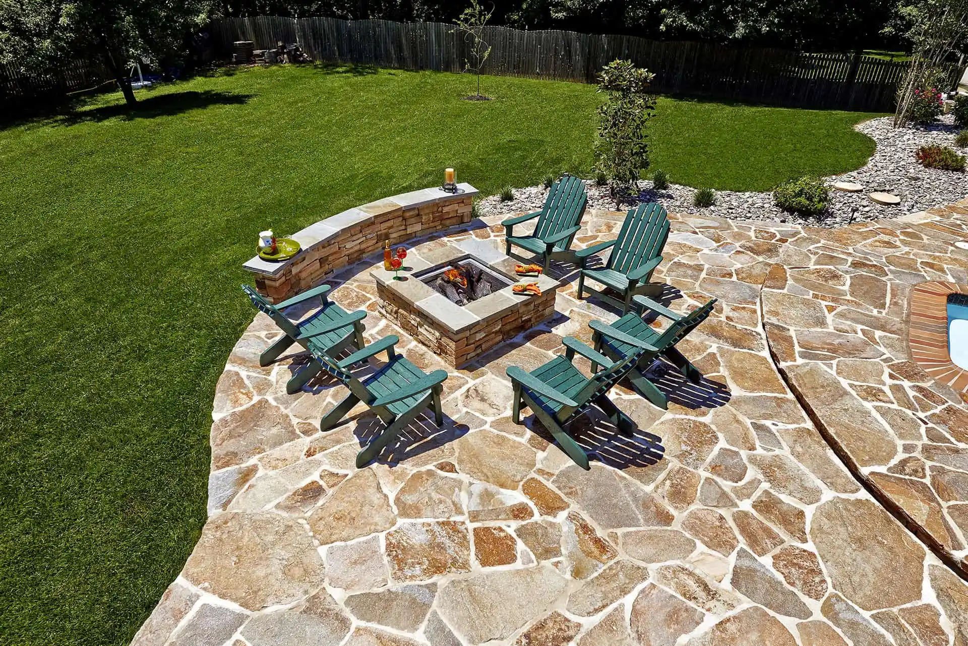 A stone patio with a square fire pit, surrounded by six green Adirondack chairs, with grassy lawn and a curved stone path visible in the background.