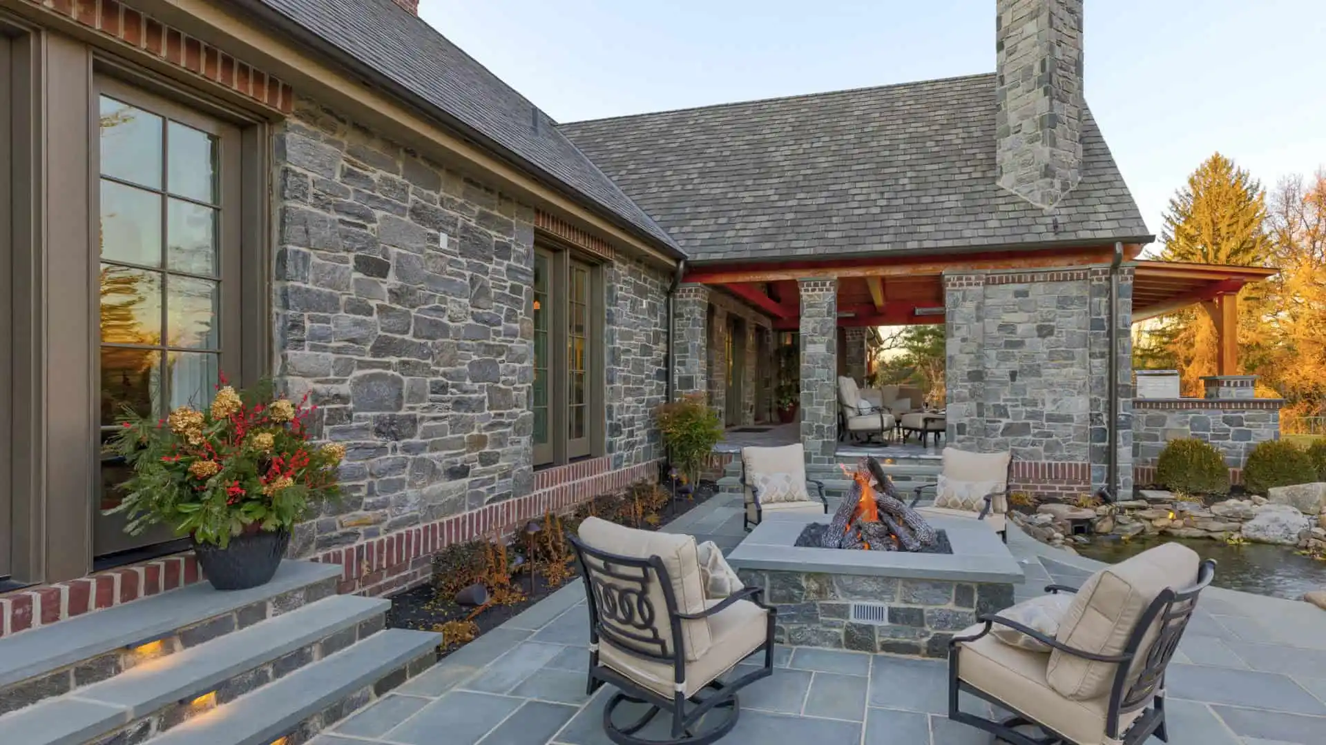 Stone house with a cozy outdoor patio featuring cushioned chairs around a square fire pit. Enjoy enhanced outdoor living in Delaware County, PA, with large windows and a shingled roof. A small pond enhances the landscaped plants near the patio, complemented by expert hardscaping services.