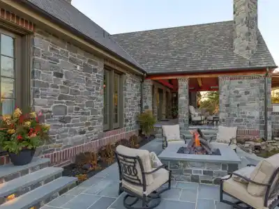 Stone house with a cozy outdoor patio featuring cushioned chairs around a square fire pit. Enjoy enhanced outdoor living in Delaware County, PA, with large windows and a shingled roof. A small pond enhances the landscaped plants near the patio, complemented by expert hardscaping services.