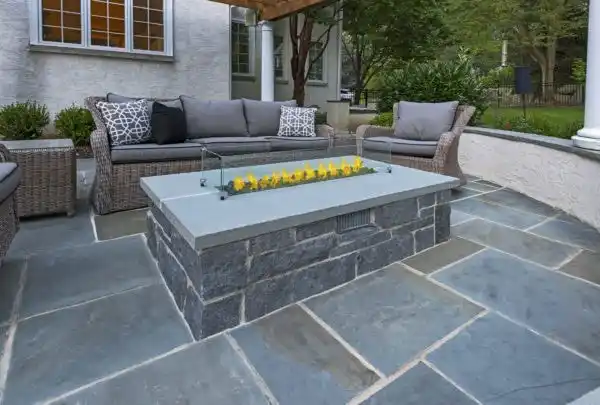 This patio in Delaware County, PA boasts gray stone flooring and elegant wicker furniture with gray cushions. A sleek rectangular fire pit adds modern flair, while lush greenery envelops the space. Perfect for outdoor living, enhanced by expert hardscaping services.