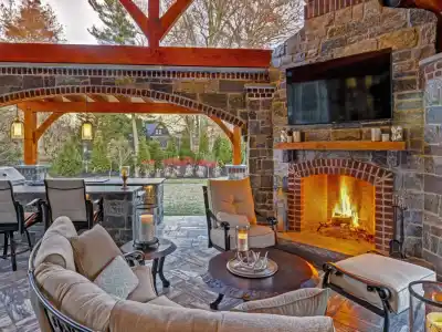 A cozy outdoor living space in Delaware County, PA features a stone fireplace with a crackling fire, cushioned seating, and a round coffee table adorned with candles. A bar area is tucked under a wooden pergola, while expert hardscaping services frame the trees and garden in the distance.