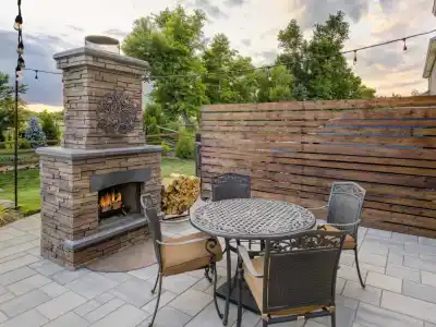 A cozy outdoor patio in Delaware County, PA, showcases captivating outdoor living with a stone fireplace, round metal table with chairs, and a wooden privacy fence. String lights dangle above the lush greenery and vibrant garden under a cloudy sky.