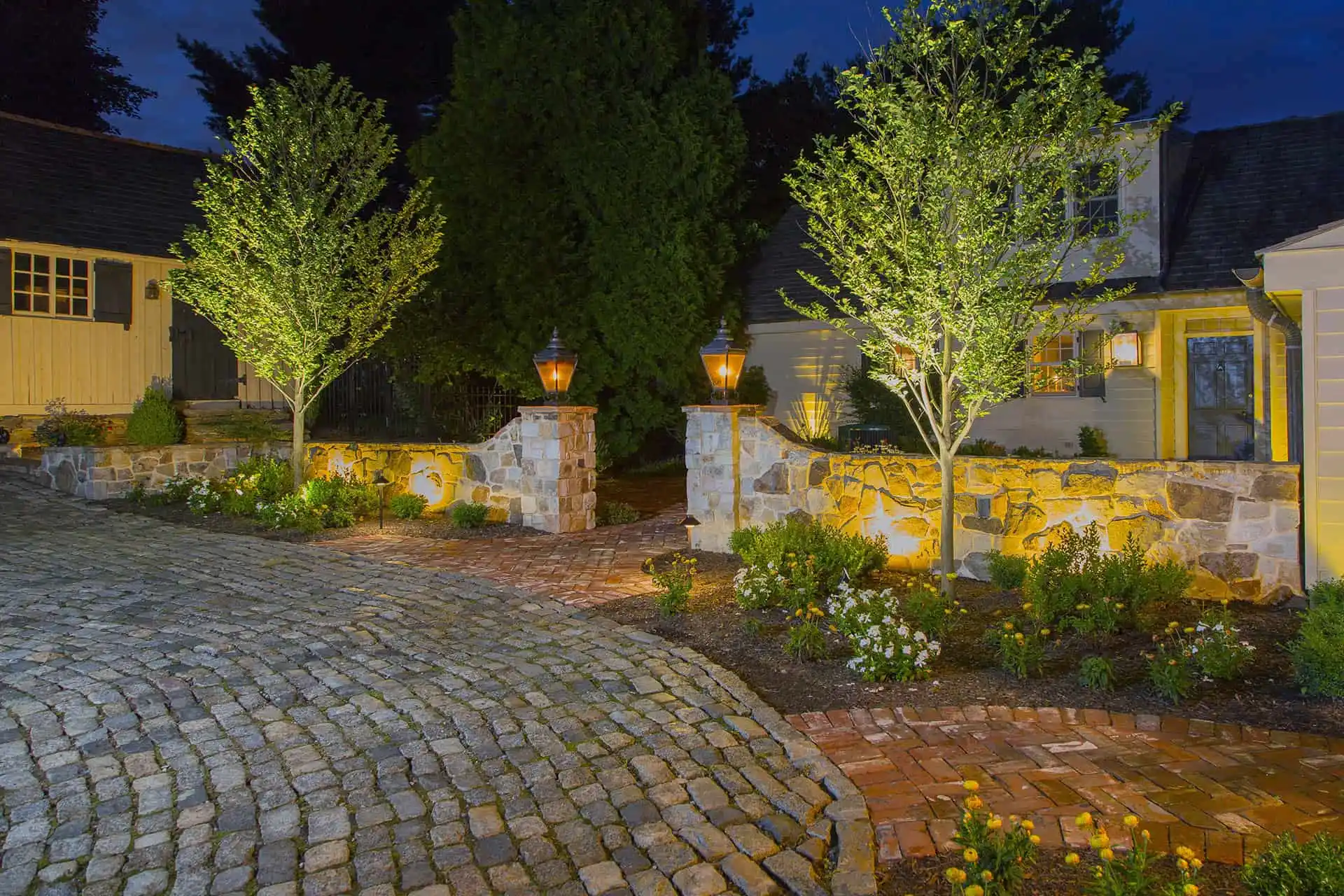 A well-lit cobblestone driveway leads to a house at night, bordered by garden beds, trees, and a stone wall with lantern lights, creating a warm and inviting entrance.