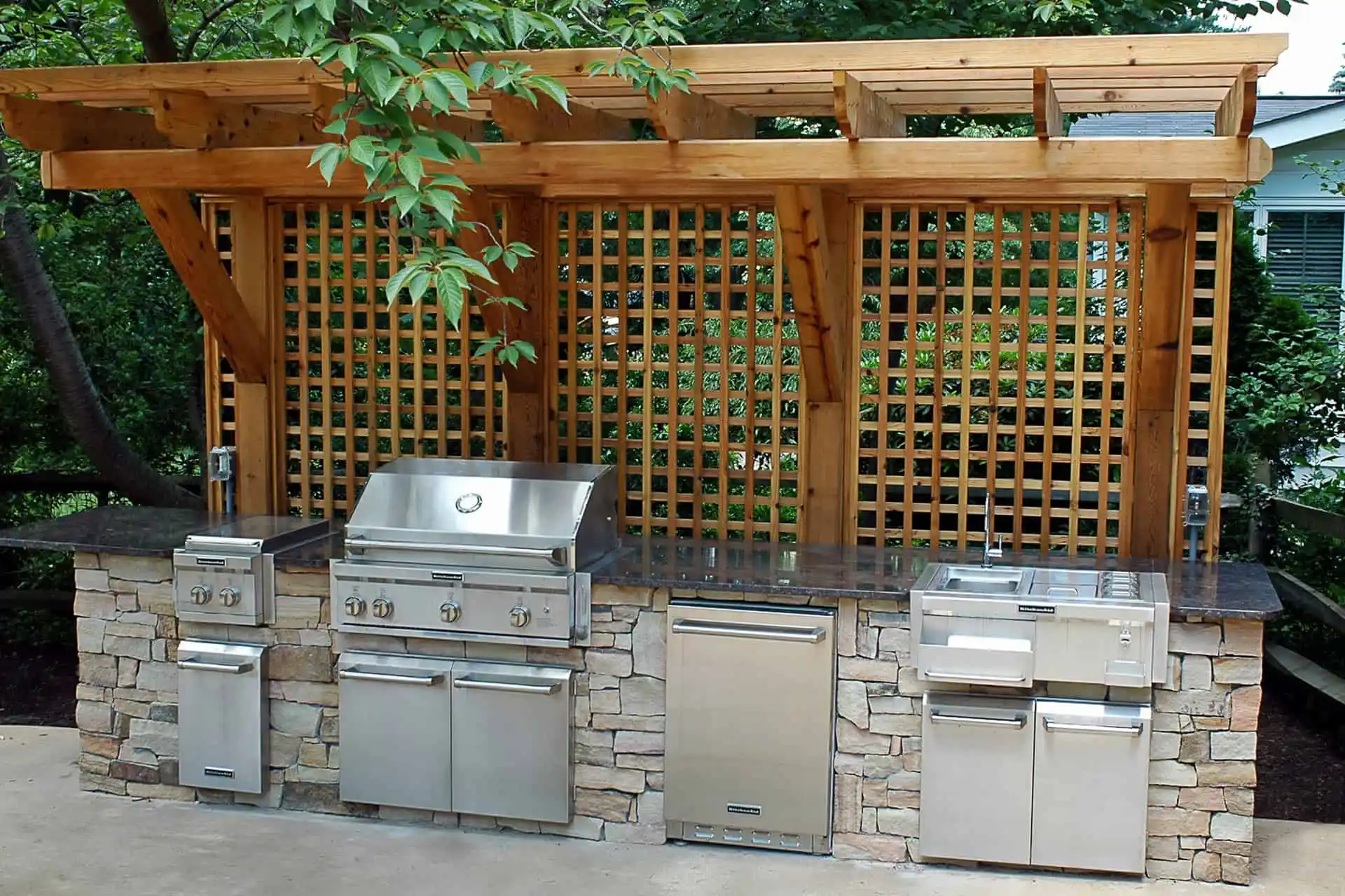 Outdoor kitchen with built-in stainless steel grill, side burner, refrigerator, and sink, set in a stone counter beneath a wooden pergola with lattice backdrop, surrounded by trees and greenery.