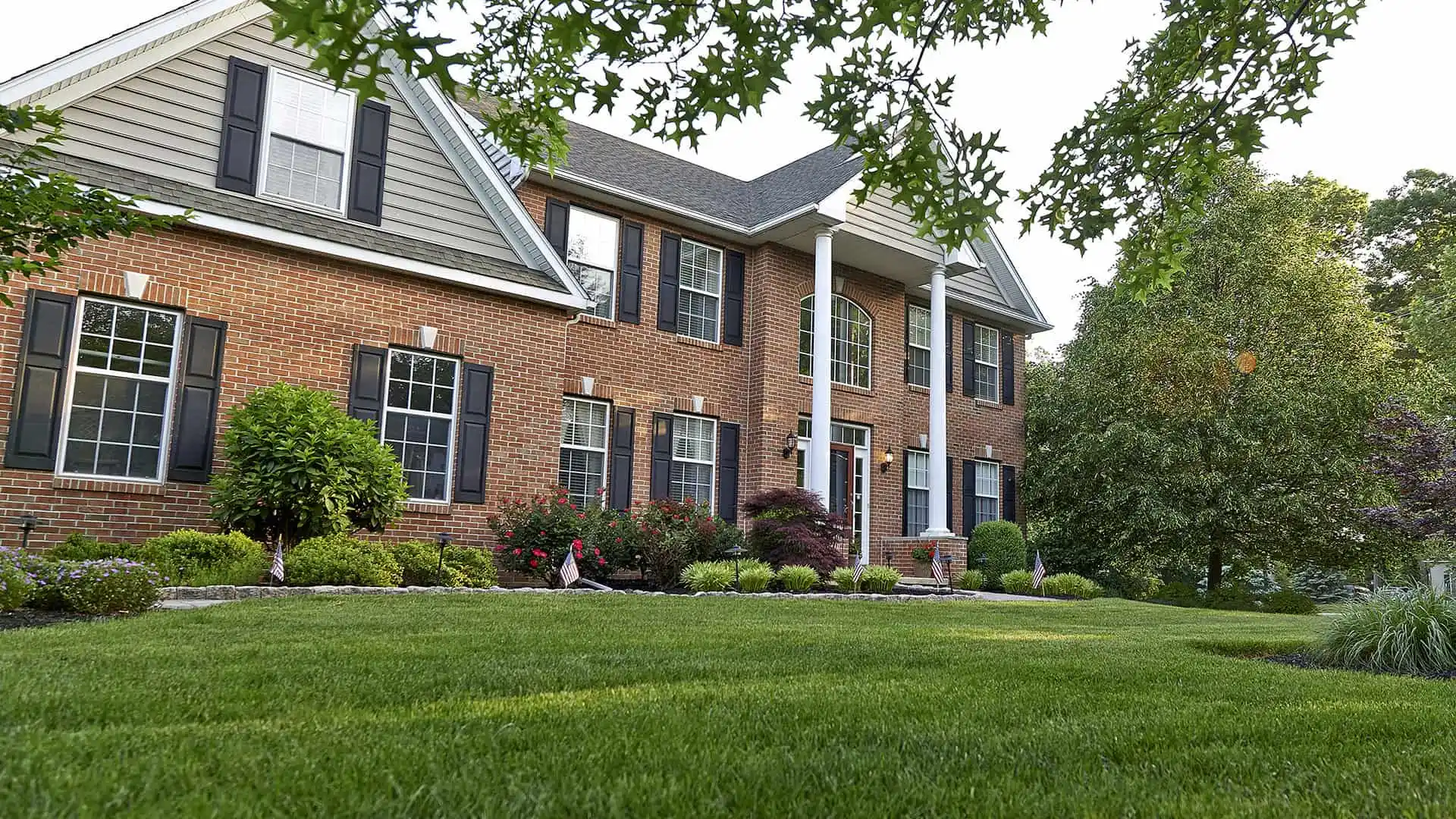 A two-story brick house with a well-maintained lawn offers ideal outdoor living in Delaware County, PA. The house features multiple windows with shutters, surrounded by neatly trimmed bushes and trees. The sky is clear, creating a serene suburban scene perfect for enjoying expert landscaping services.