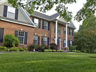 A two-story brick house with a well-maintained lawn offers ideal outdoor living in Delaware County, PA. The house features multiple windows with shutters, surrounded by neatly trimmed bushes and trees. The sky is clear, creating a serene suburban scene perfect for enjoying expert landscaping services.