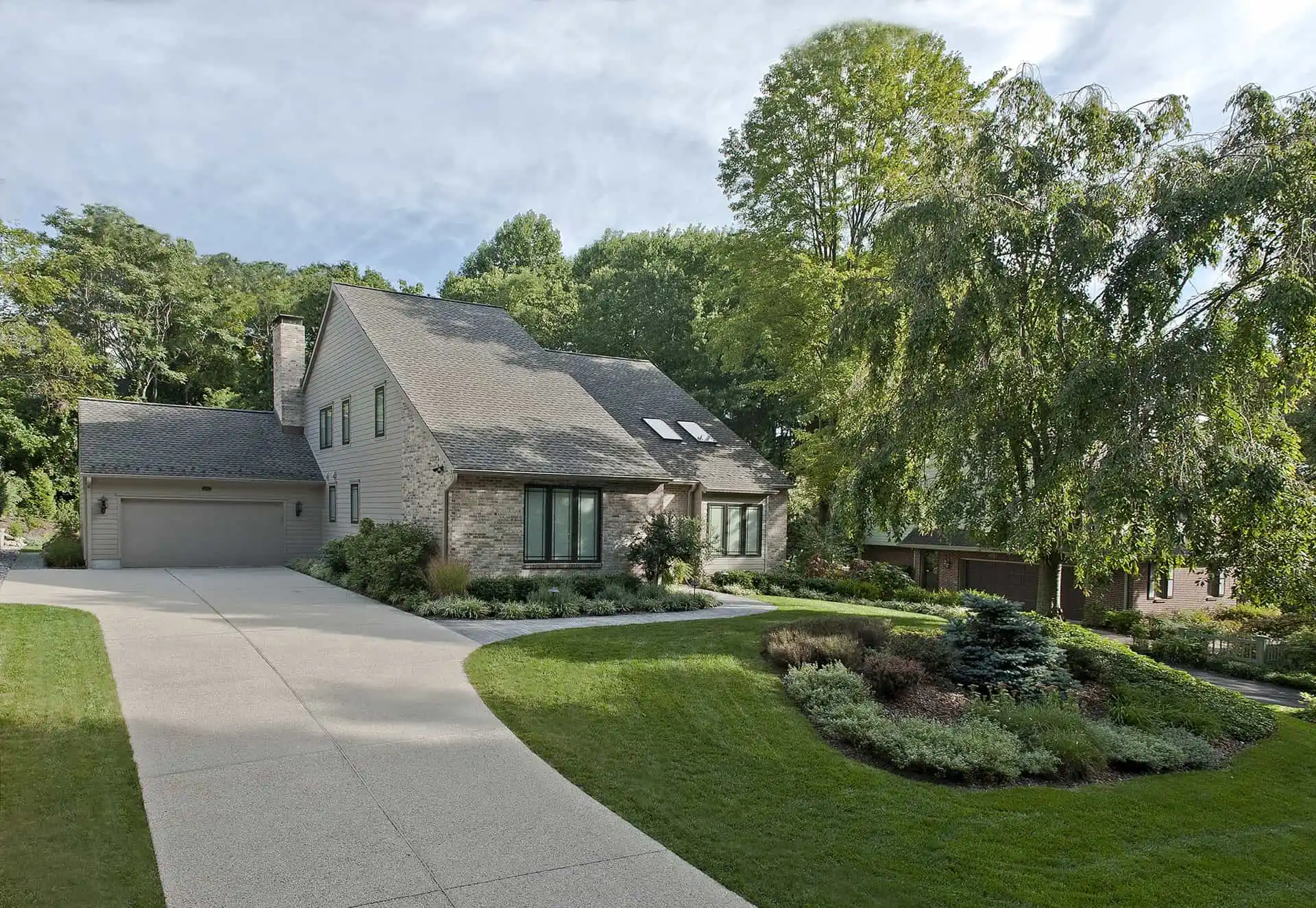 A modern two-story house with a sloped roof, brick and siding exterior, attached garage, and well-maintained landscaping, including a curved driveway and green lawn with trees and shrubs.