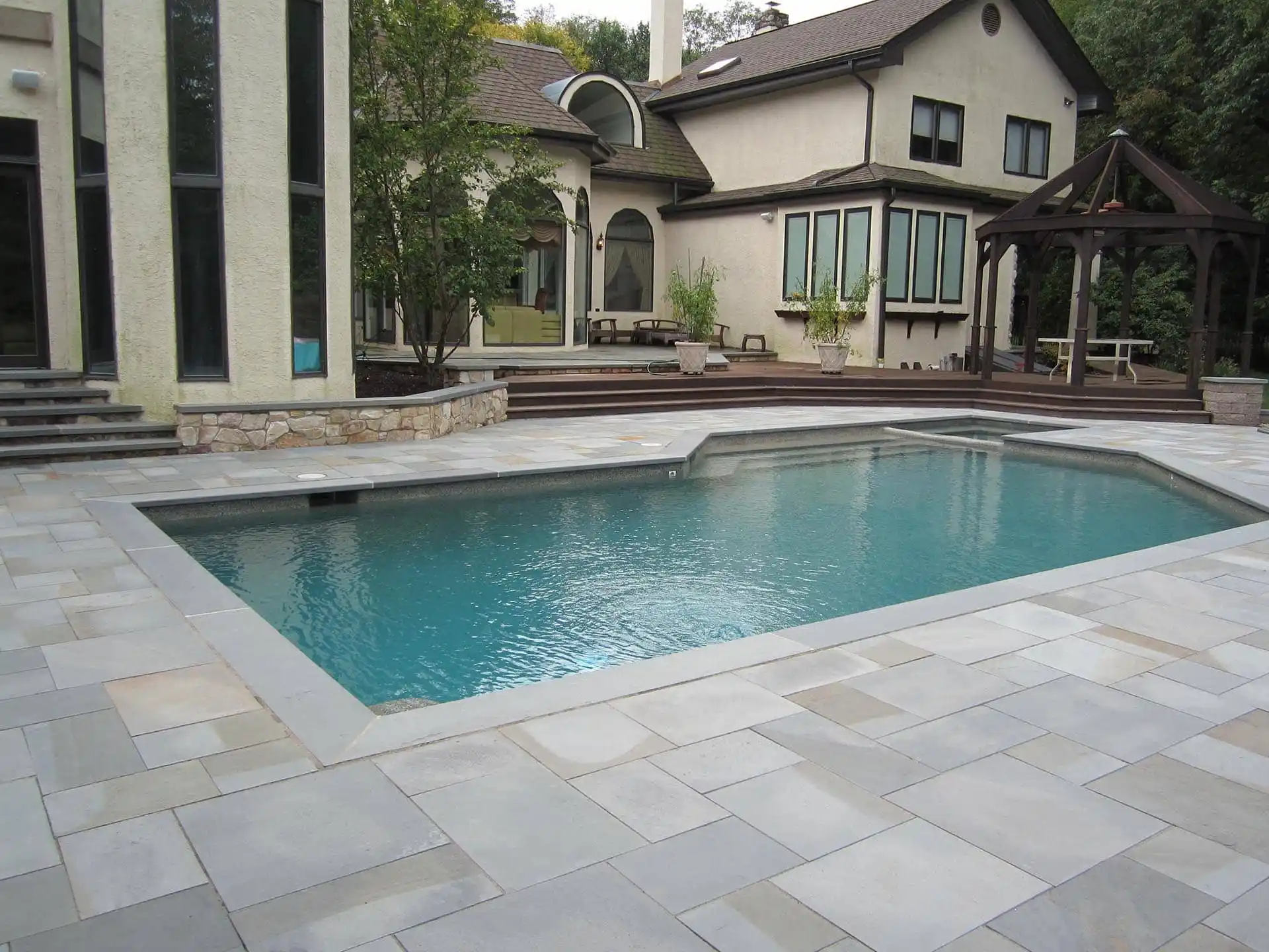 A modern backyard with a rectangular swimming pool surrounded by light gray stone tiles, a wooden deck, trees, and a large beige house with many windows and a covered patio area.