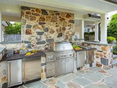 Outdoor kitchen with a stone facade featuring a stainless steel grill, refrigerator, and cabinets. The countertop holds lemons and a tray of herbs. Covered by a white ceiling and surrounded by greenery, this space reflects expert outdoor living design in Delaware County, PA.