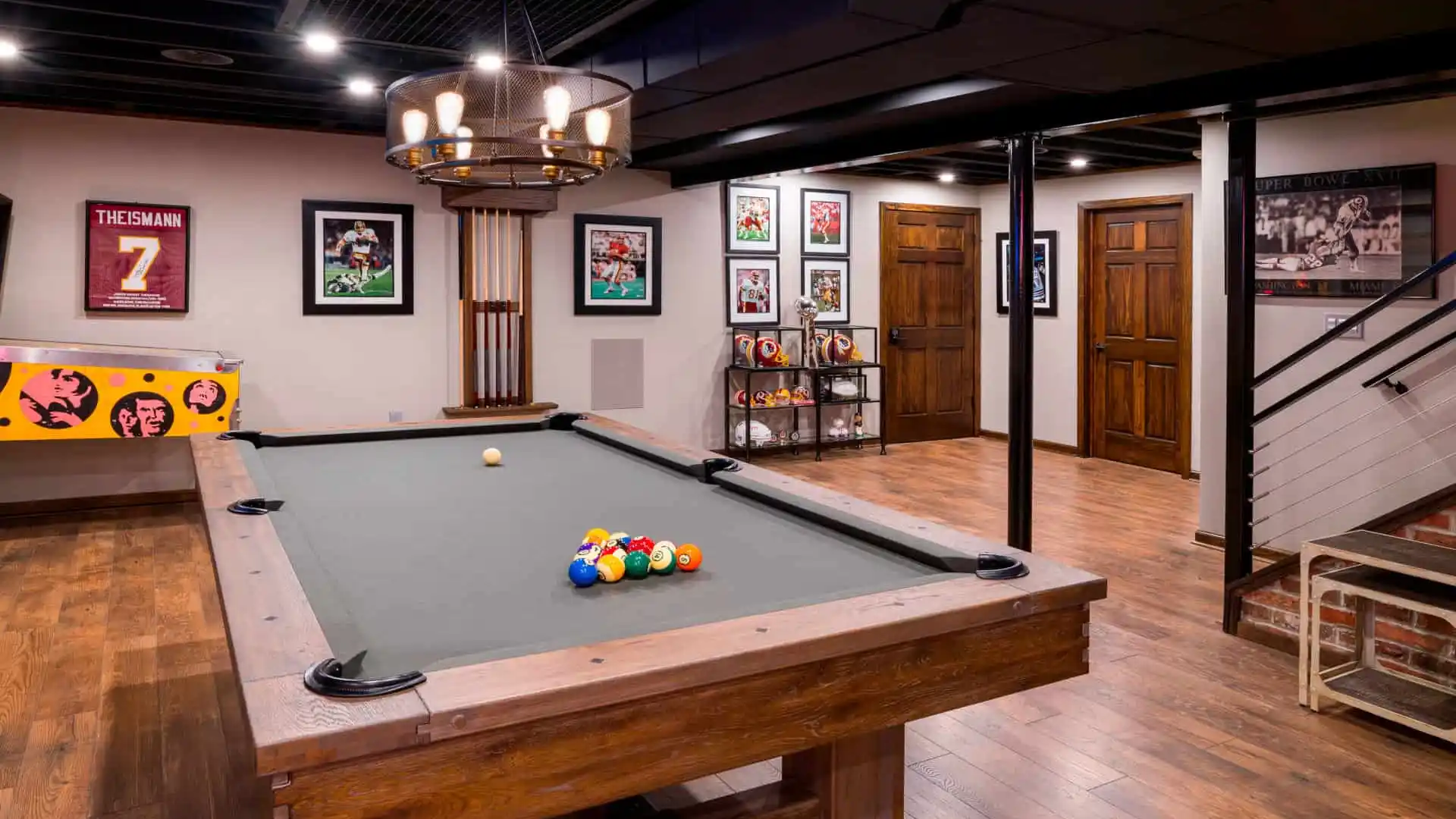 A basement game room showcases a wooden pool table with balls perfectly set up. Sports memorabilia, framed jerseys, and photos adorn the walls. A classic pinball machine sits in the corner, enhancing this cozy hideaway. Experience expert hardscaping services in Delaware County, PA to transform your space.