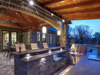 Outdoor living in Delaware County, PA comes to life with a kitchen featuring a stone and wood patio, built-in grill, countertop bar seating, and ambient lighting. A dining table and seating area sit under the twilight sky, surrounded by trees for the perfect outdoor escape.