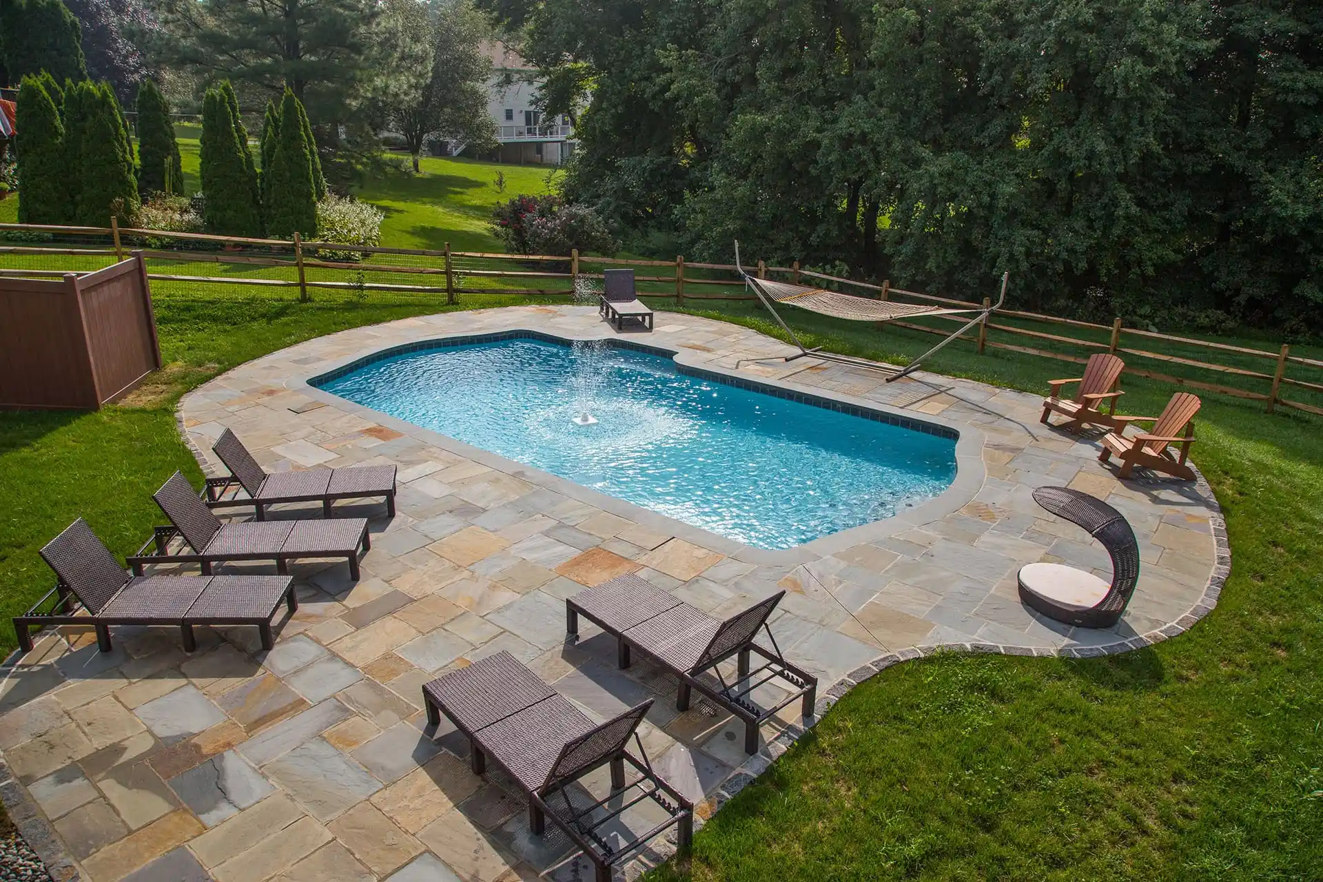 An outdoor swimming pool surrounded by stone patio, several lounge chairs, a hammock, a wooden fence, and trees; green lawn and landscaped garden visible in the background.