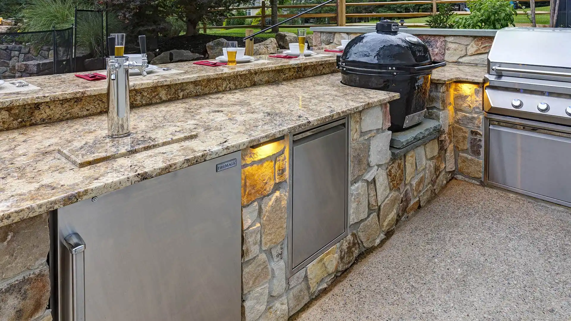 Experience outdoor living in Delaware County, PA, with this outdoor kitchen. It boasts a stone counter accented by a granite top, featuring a grill, black Kamado-style smoker, and keg tap. Red placemats with glasses of beer sit atop the sleek stone base and its convenient small cabinets below.