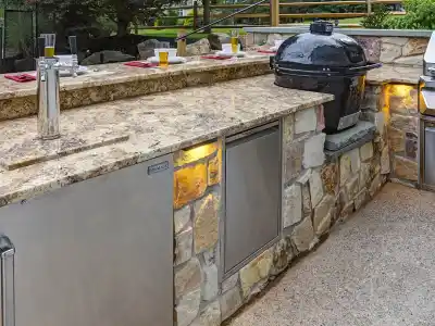 Experience outdoor living in Delaware County, PA, with this outdoor kitchen. It boasts a stone counter accented by a granite top, featuring a grill, black Kamado-style smoker, and keg tap. Red placemats with glasses of beer sit atop the sleek stone base and its convenient small cabinets below.