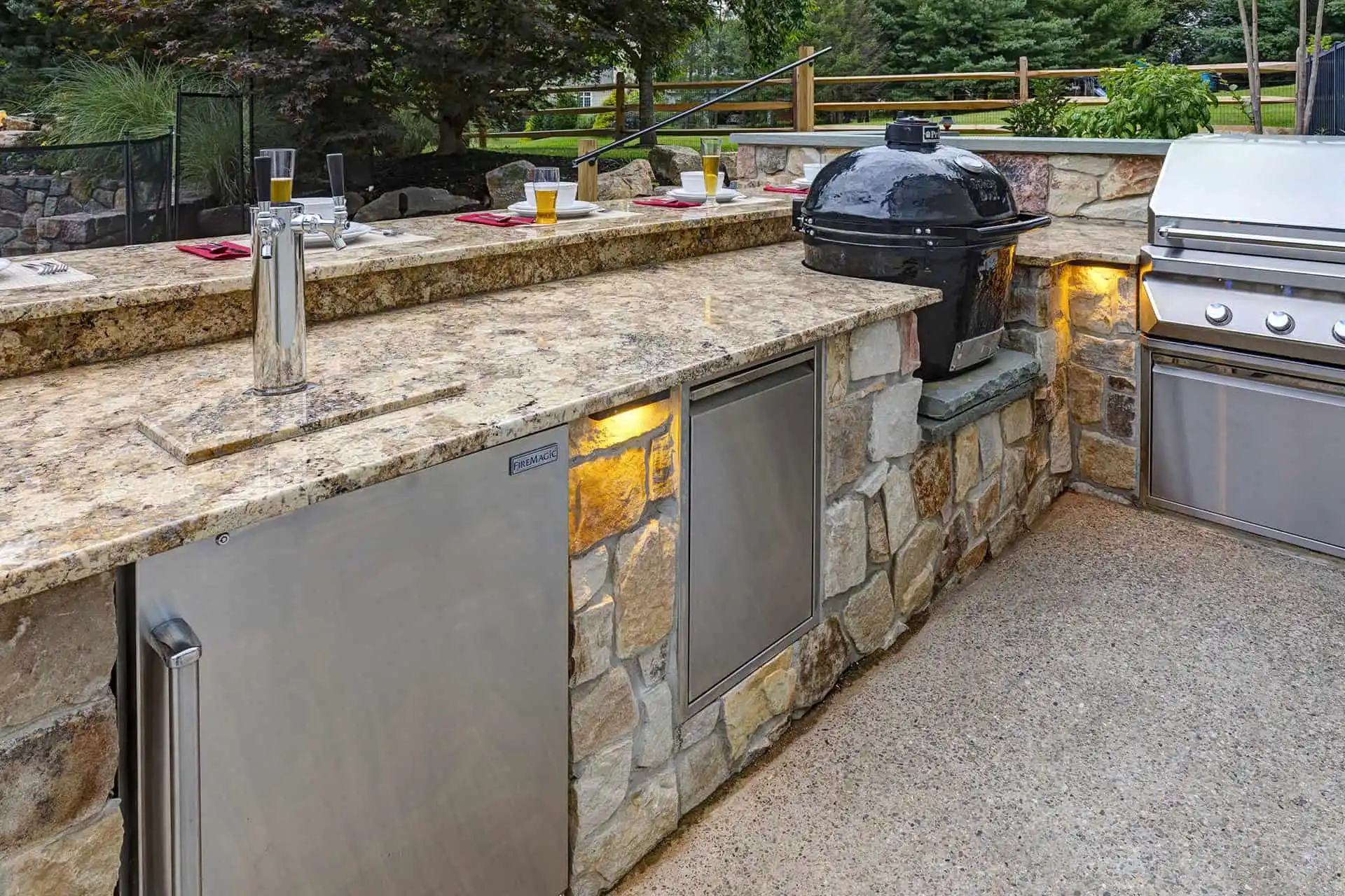 Outdoor kitchen with stone countertops, built-in stainless steel fridge, grill, and a black ceramic kamado-style smoker. Four glasses with drinks and red napkins are placed on the counter. Trees and a fence are in the background.
