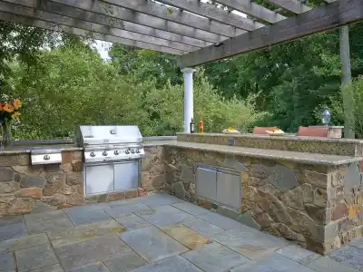 Outdoor living in Delaware County, PA is elevated by this kitchen under a wooden pergola featuring a built-in grill, stone countertops, and bar stools. Fresh flowers and condiments adorn the surface. Enveloped by trees and a stone patio, it's crafted with expert hardscaping services in Delaware County, PA.