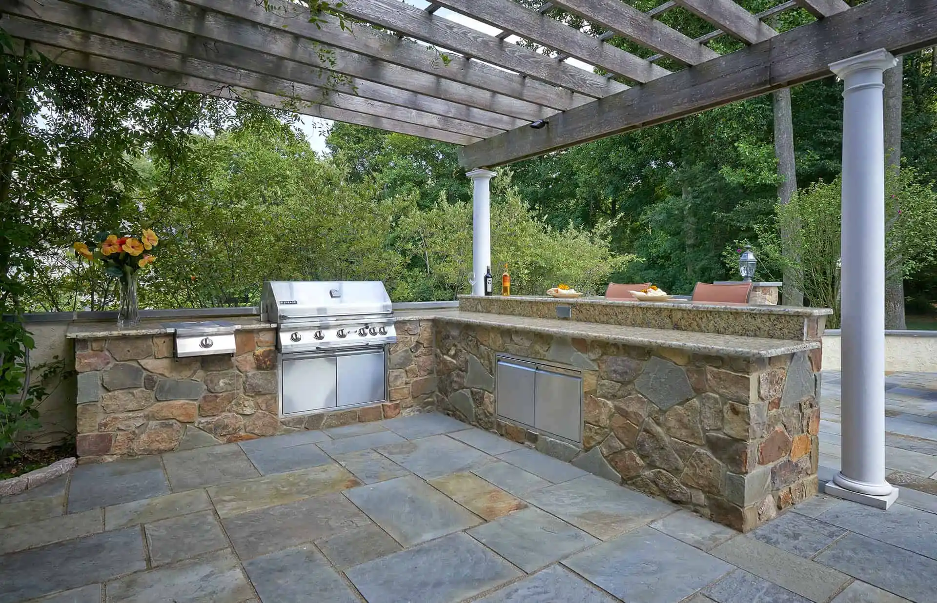 Outdoor kitchen with a built-in grill, stone countertops, pergola overhead, and bar seating. Surrounded by trees and greenery, with a vase of flowers on the counter and patio flooring.