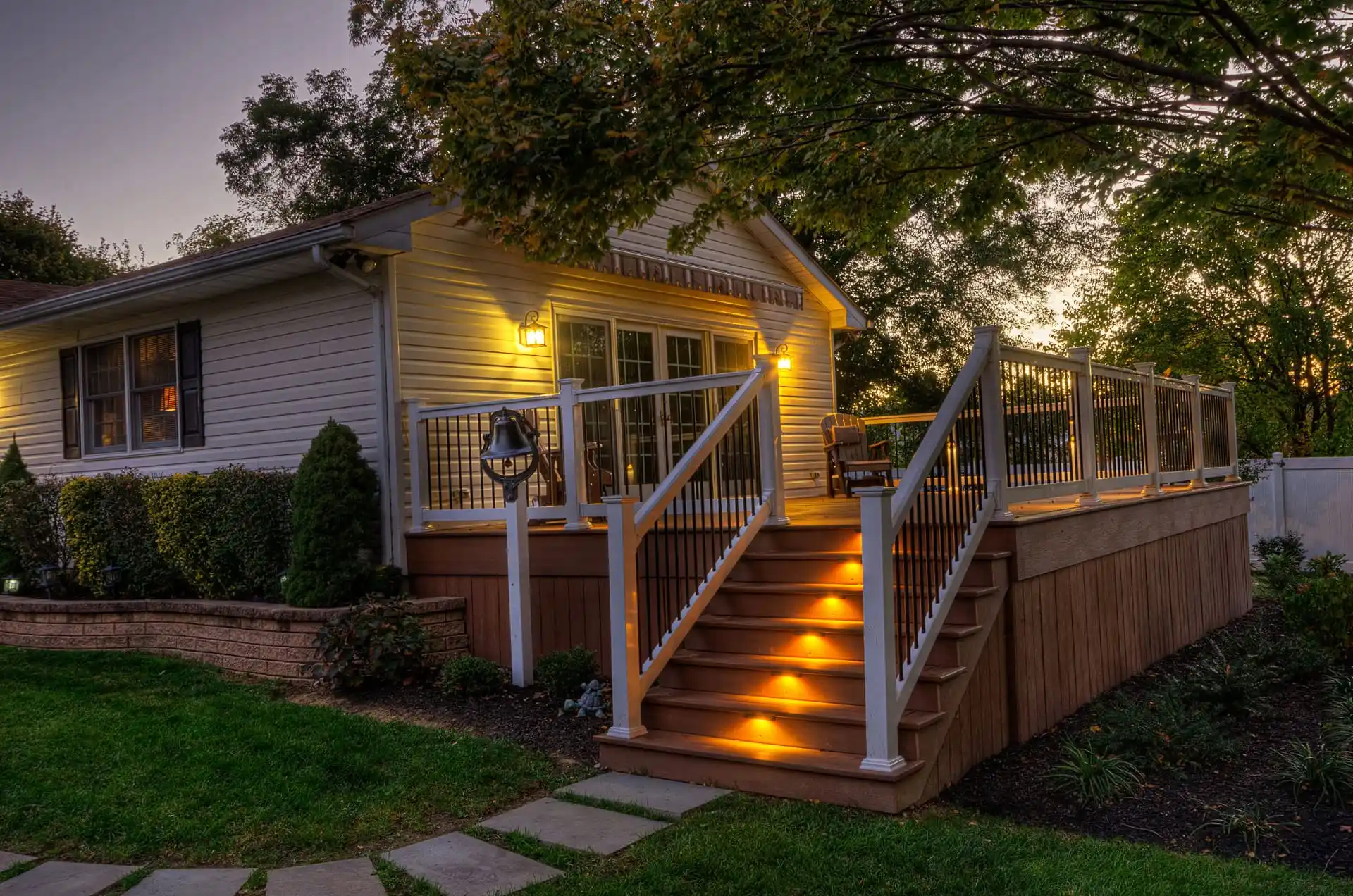 A cozy house with a large wooden deck and illuminated stair lights at dusk, surrounded by green grass, shrubs, and trees. The deck has a white railing and outdoor chairs, creating a warm, inviting atmosphere.