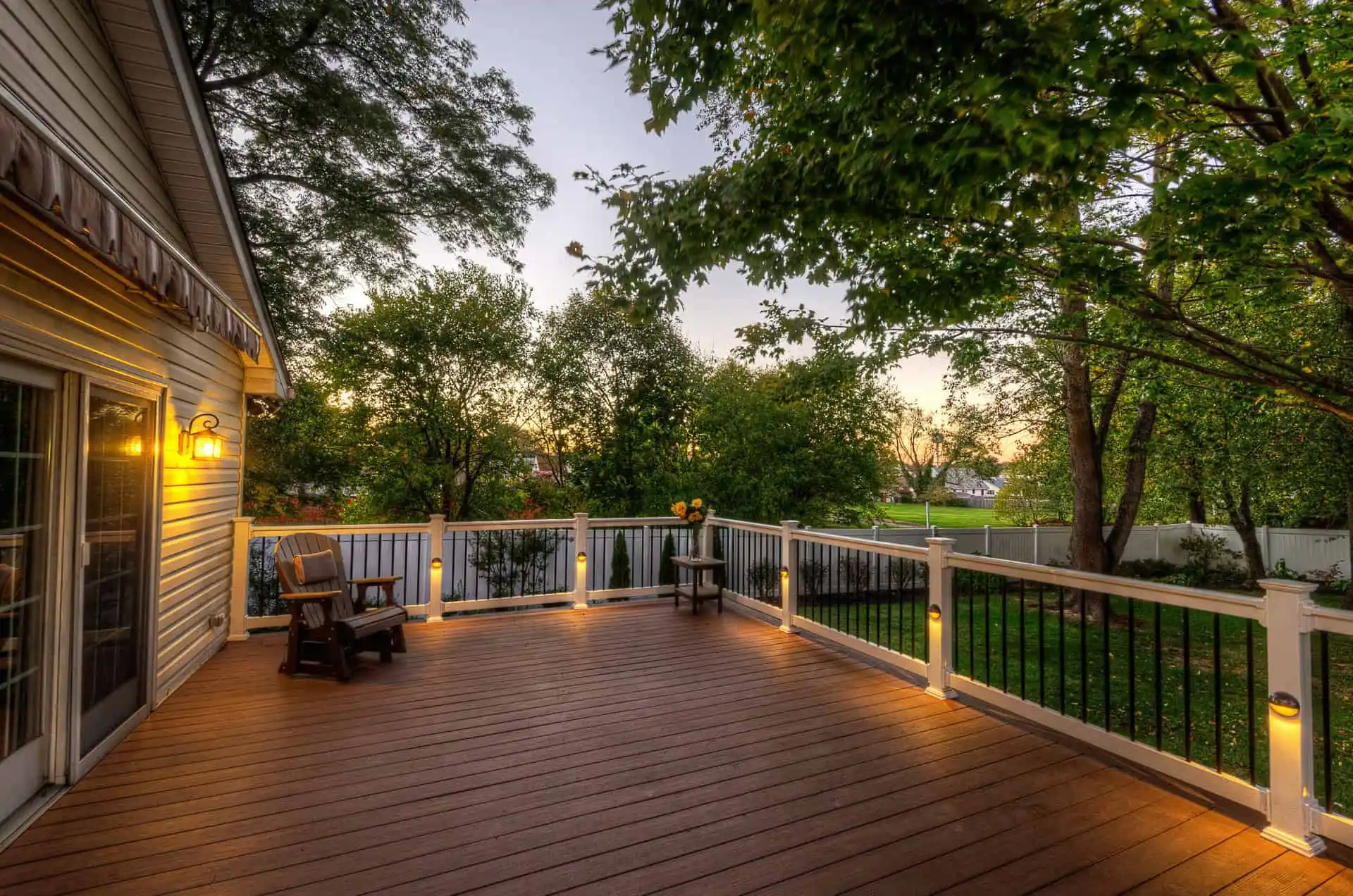 Spacious wooden deck with railing, outdoor lighting, and a few chairs, overlooking a fenced backyard with trees at sunset. Warm lights create a cozy, inviting atmosphere.