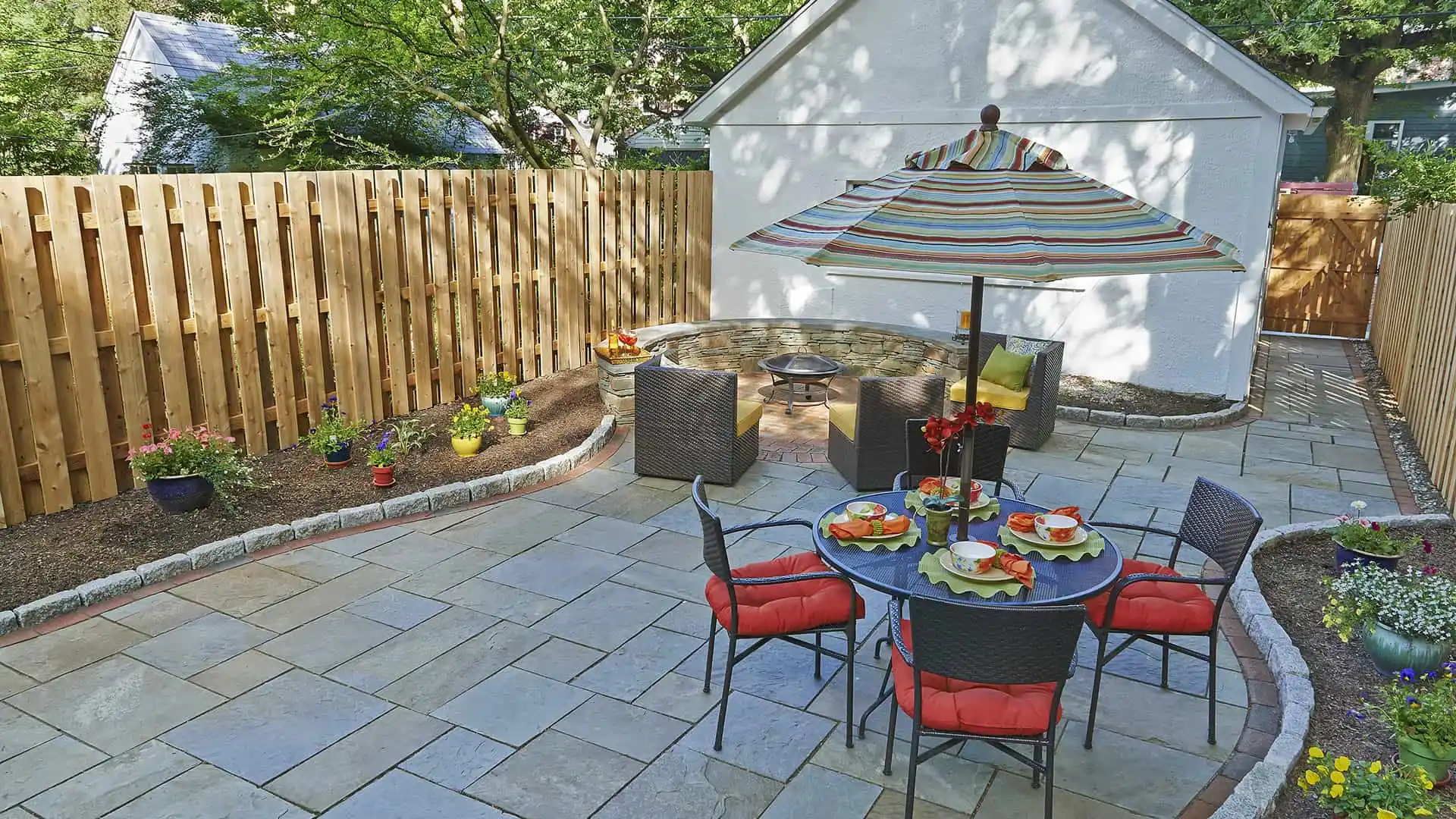 The backyard patio, a masterpiece of outdoor living in Delaware County, PA, features a stone fire pit with cozy wicker chairs, a dining table set with vibrant plates beneath a striped umbrella. Enveloped by lush flowers and plants, its charm is enhanced by expert landscaping services.