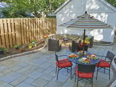 The backyard patio, a masterpiece of outdoor living in Delaware County, PA, features a stone fire pit with cozy wicker chairs, a dining table set with vibrant plates beneath a striped umbrella. Enveloped by lush flowers and plants, its charm is enhanced by expert landscaping services.