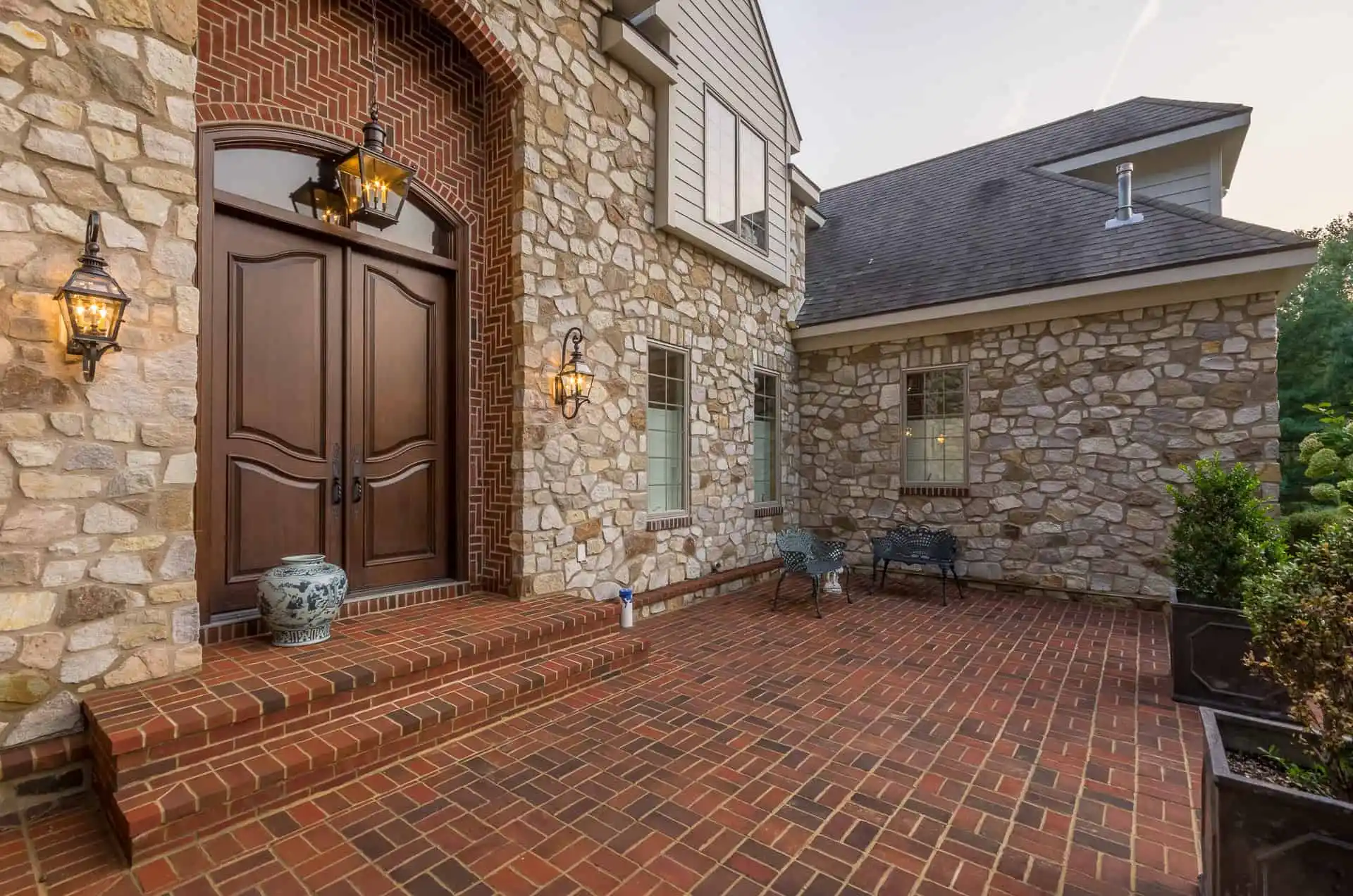 A brick patio with decorative lanterns and stone walls leads to large double wooden doors at the entrance of a house. Patio furniture and potted plants are arranged along the side.