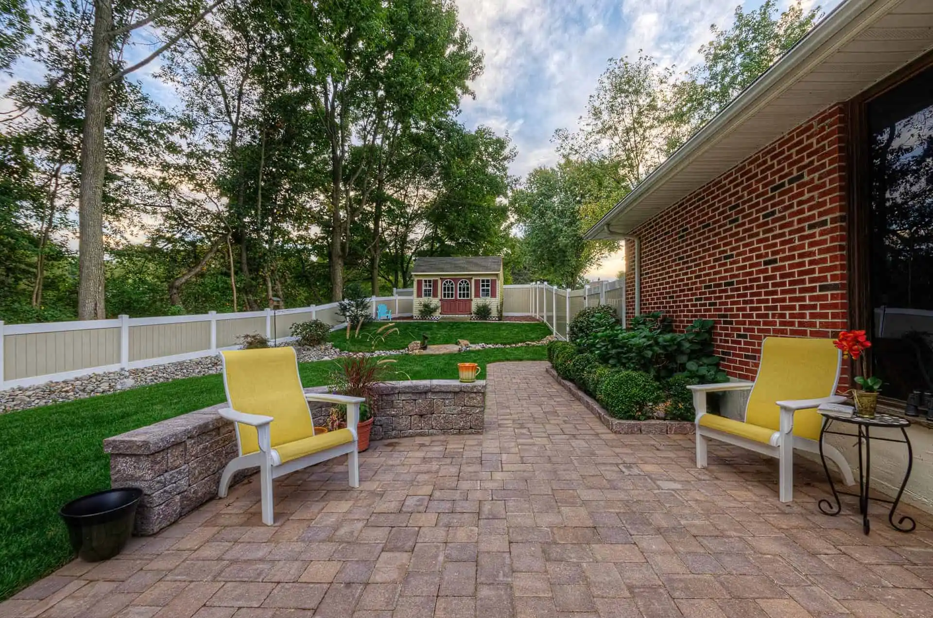 A backyard patio with two yellow chairs, stone planters, potted plants, and a brick pathway leading to a small shed. The area is surrounded by a white fence and lush green grass with trees in the background.