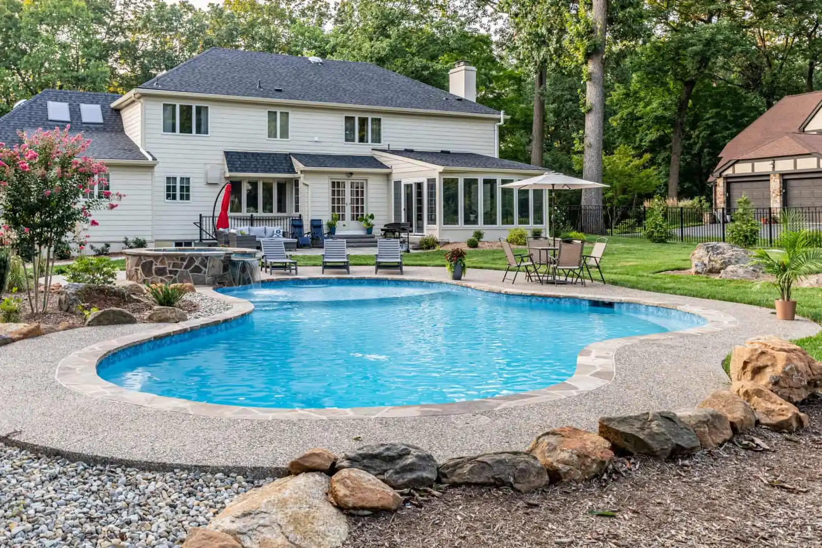 Backyard view of a white two-story house with a patio, surrounded by trees. A kidney-shaped swimming pool is in the foreground, bordered by rocks and chairs. Enjoy outdoor living in Delaware County, PA, with umbrellas and lounge chairs set up for relaxation nearby.