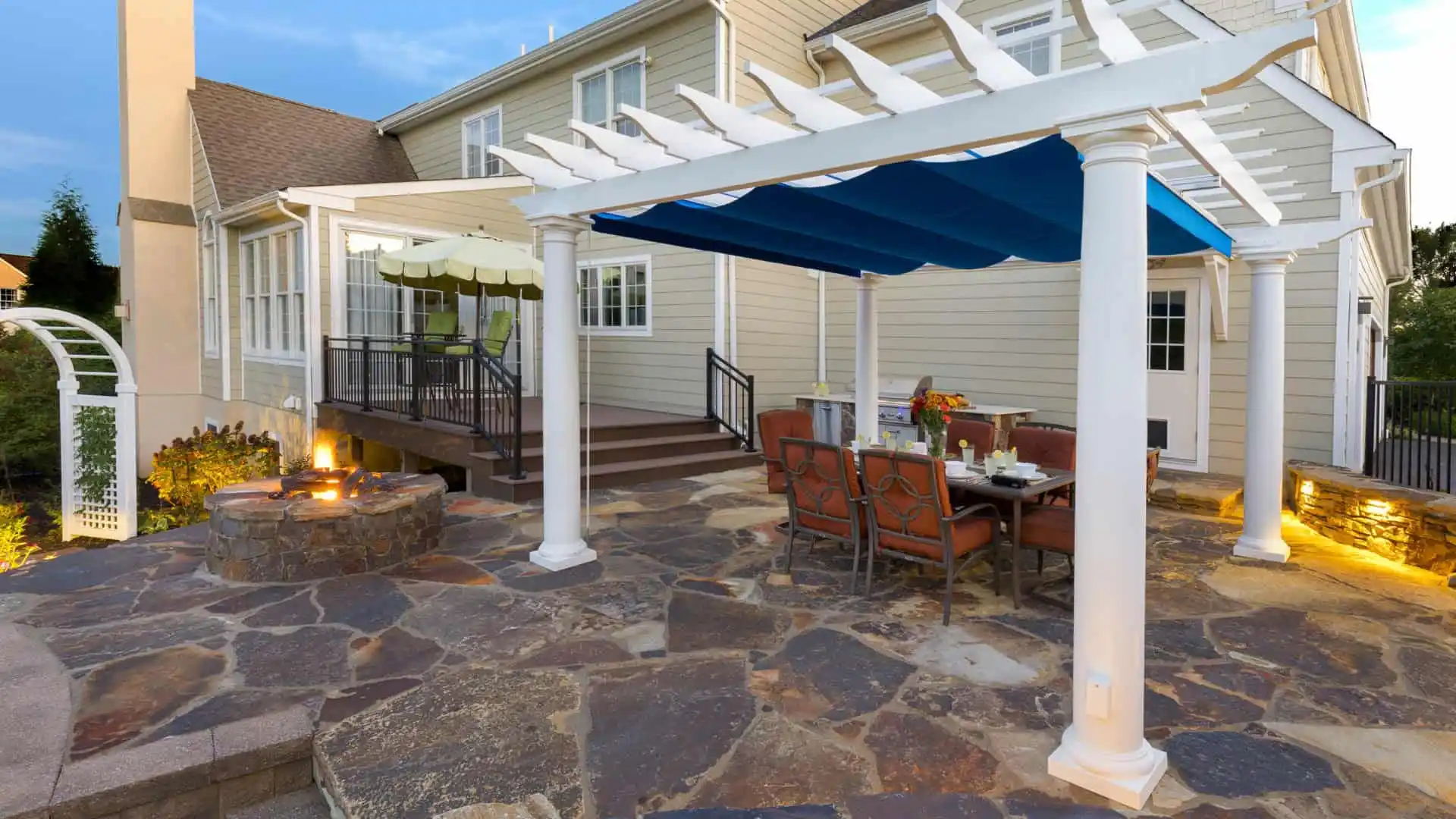 Outdoor living in Delaware County, PA, is exemplified by this patio with a pergola draped in blue fabric. Below are cushioned chairs and a table adorned with flowers. Nearby, a stone fire pit rests on stone flooring, enhancing the light-colored siding and large windows of the home.