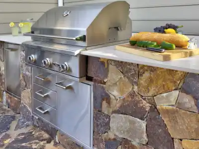 Outdoor kitchen with a stone base and stainless steel grill exemplifies outdoor living in Delaware County, PA. The countertop holds a cutting board with cucumbers, a baguette, cheese, and grapes. Nearby, a pitcher of lemonade is ready to enjoy on the beautifully crafted stone patio.
