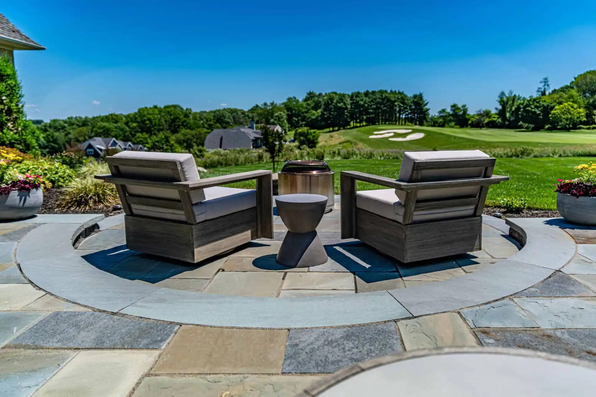 Two cushioned wooden chairs face a small round table on a circular stone patio, overlooking a scenic golf course. Bright green grass and a clear blue sky complete the tranquil setting, enhanced by expertly designed hardscaping services in Delaware County, PA, and surrounded by potted plants.