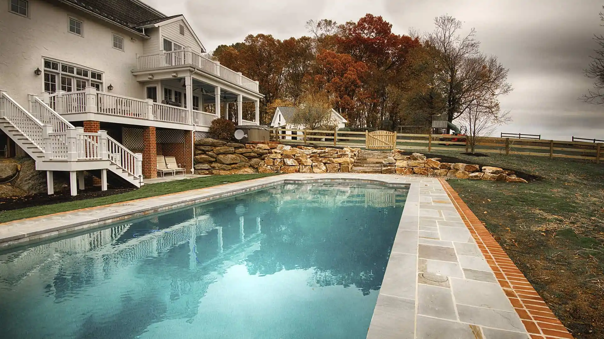A backyard swimming pool with clear blue water beside a white house with a patio and staircase, surrounded by a stone wall, autumn trees, and a wooden fence under a cloudy sky.